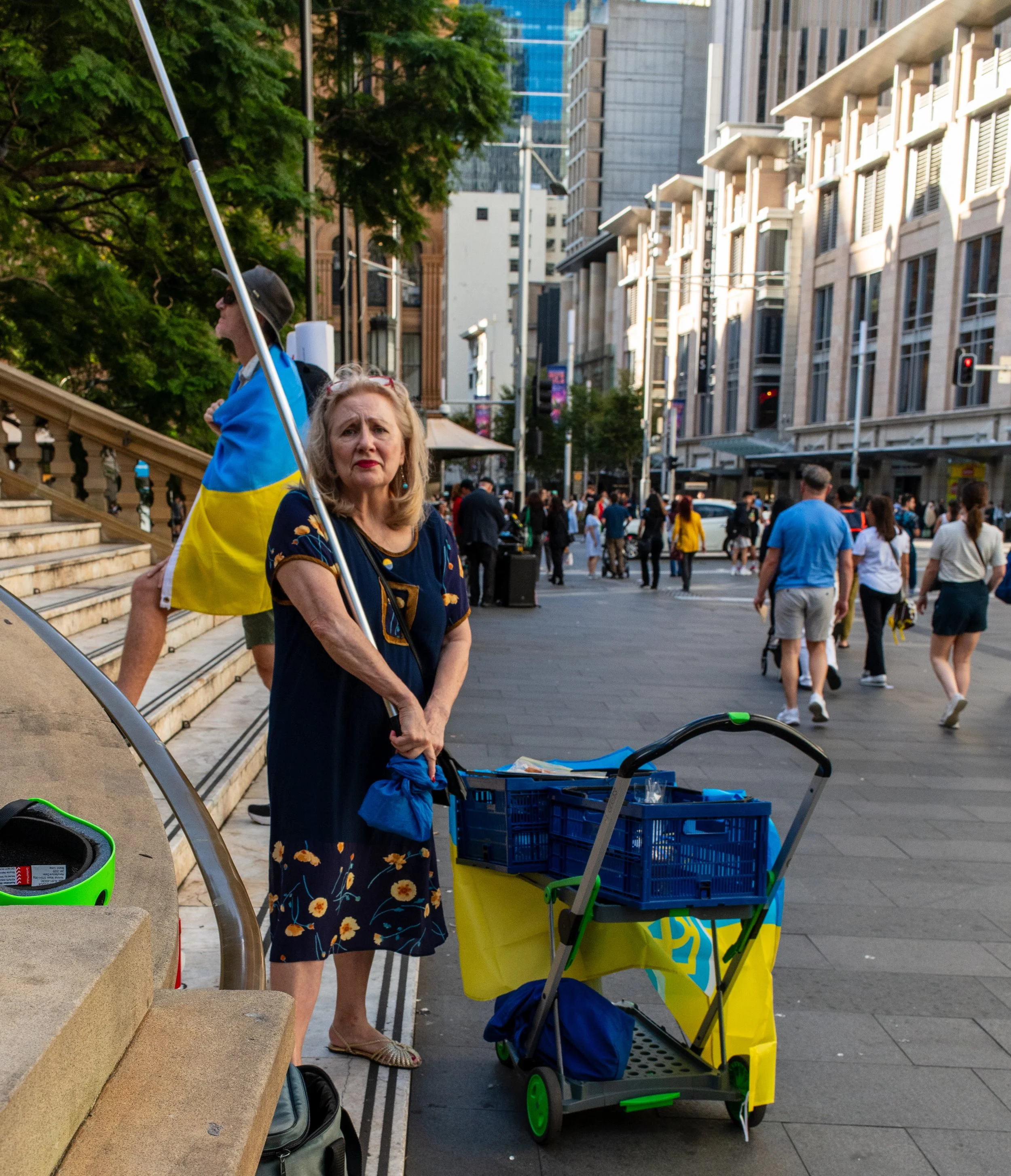 An older woman standing on a city sidewalk holding a flag of Ukraine. She is beside a shopping cart with merchandise and a black and green helmet. There are many pedestrians in the background, and tall buildings line the street.