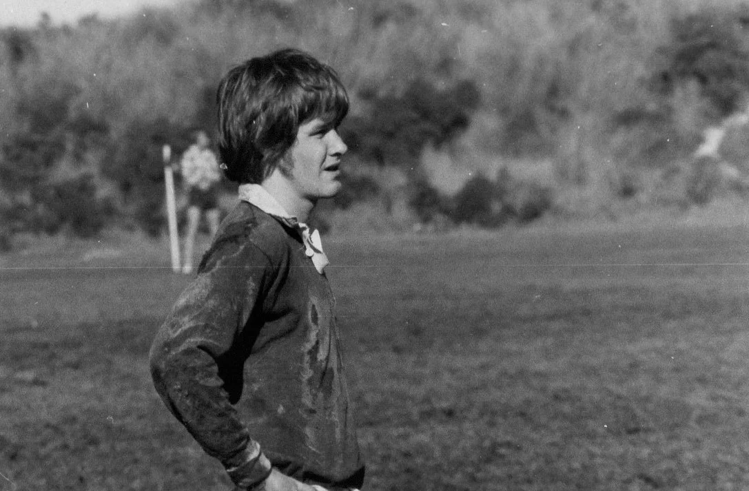 A woman wearing a jacket and a neckerchief standing outdoors on a field with trees in the background.
