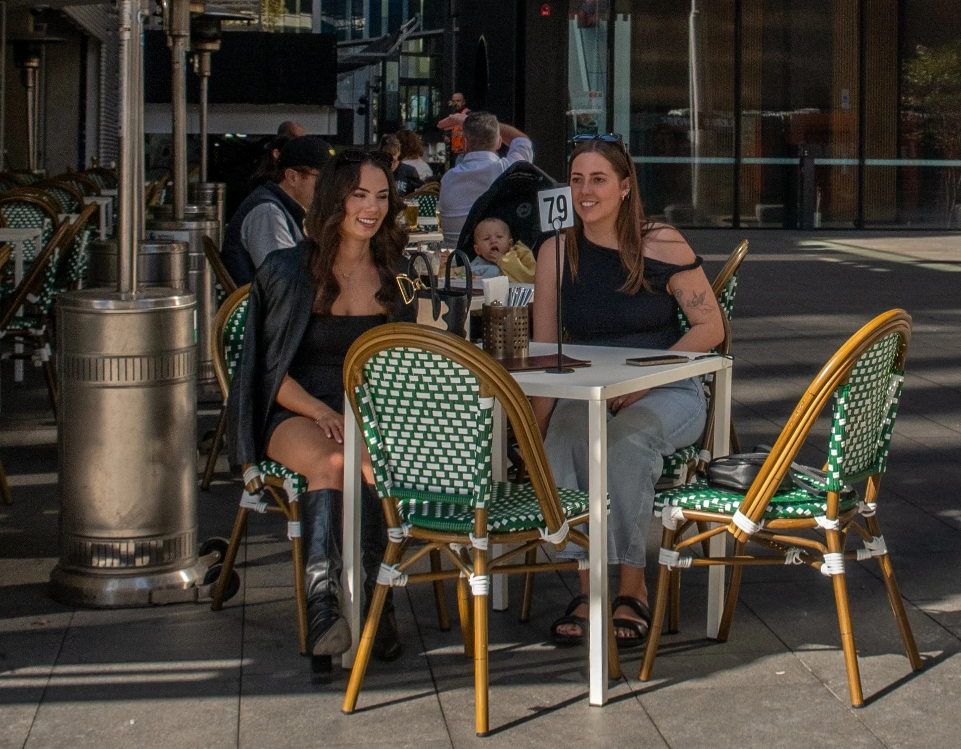 Two women sitting at an outdoor restaurant table, smiling and talking, with a baby in a stroller behind them.