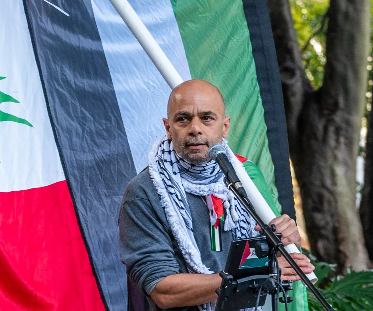 A man with a beard and bald head giving a speech at a rally or event, standing in front of a partially visible flag with black, white, green, and red colors. He is holding a microphone and has a media device in front of him, wearing a dark shirt and a keffiyeh scarf around his neck.
