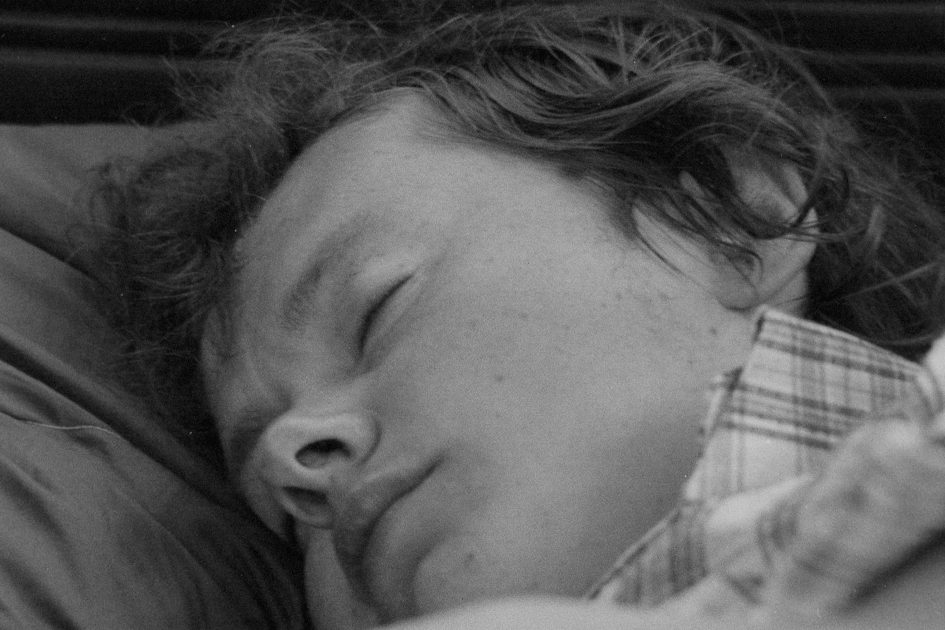 A young boy with curly hair sleeping peacefully with his face resting on a pillow, partially covered by a checkered blanket.