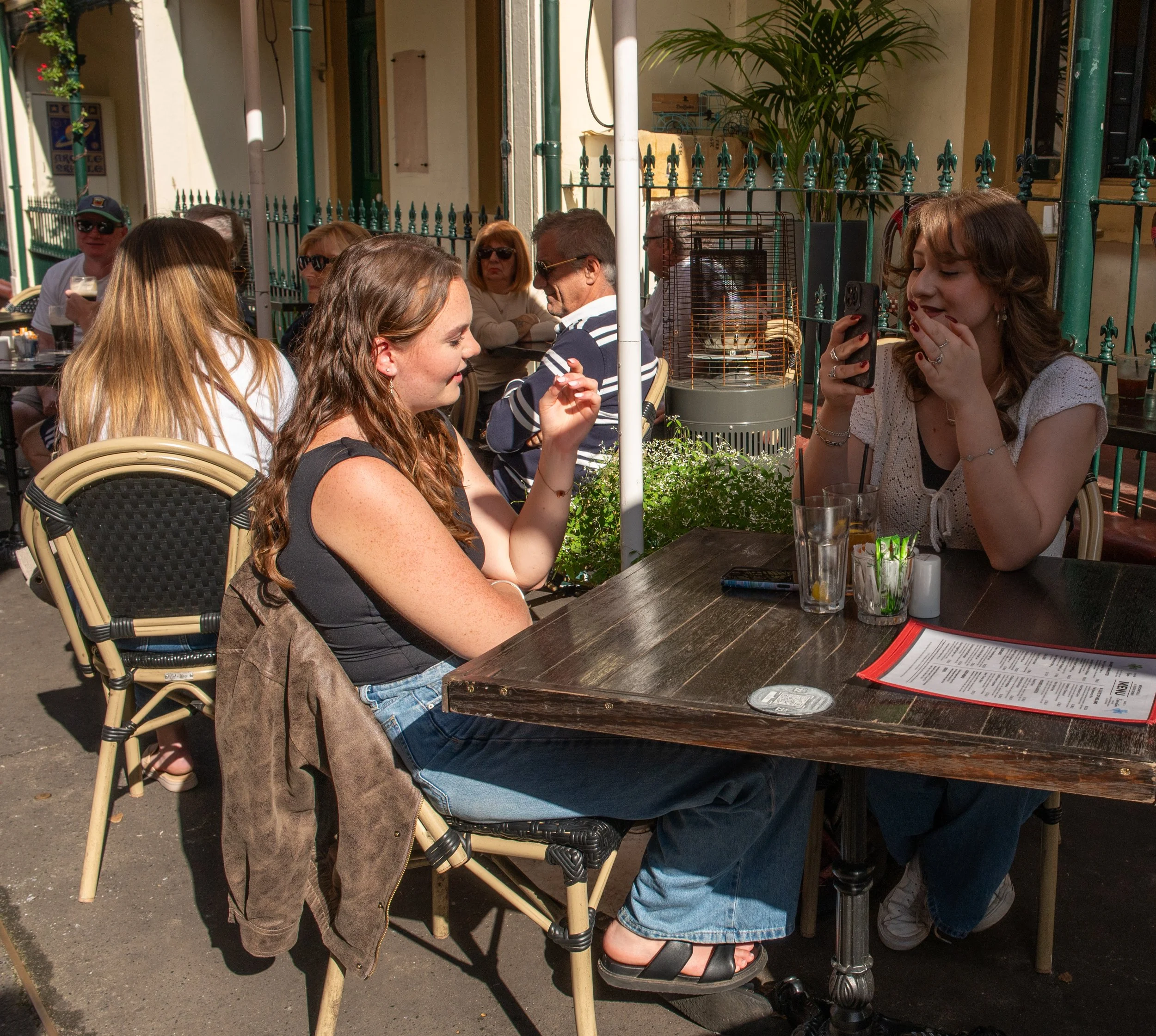Two women sit at a wooden table outside a café, one taking a photo of the other. Several people are seated in the background, enjoying drinks, with plants and a fence behind them.