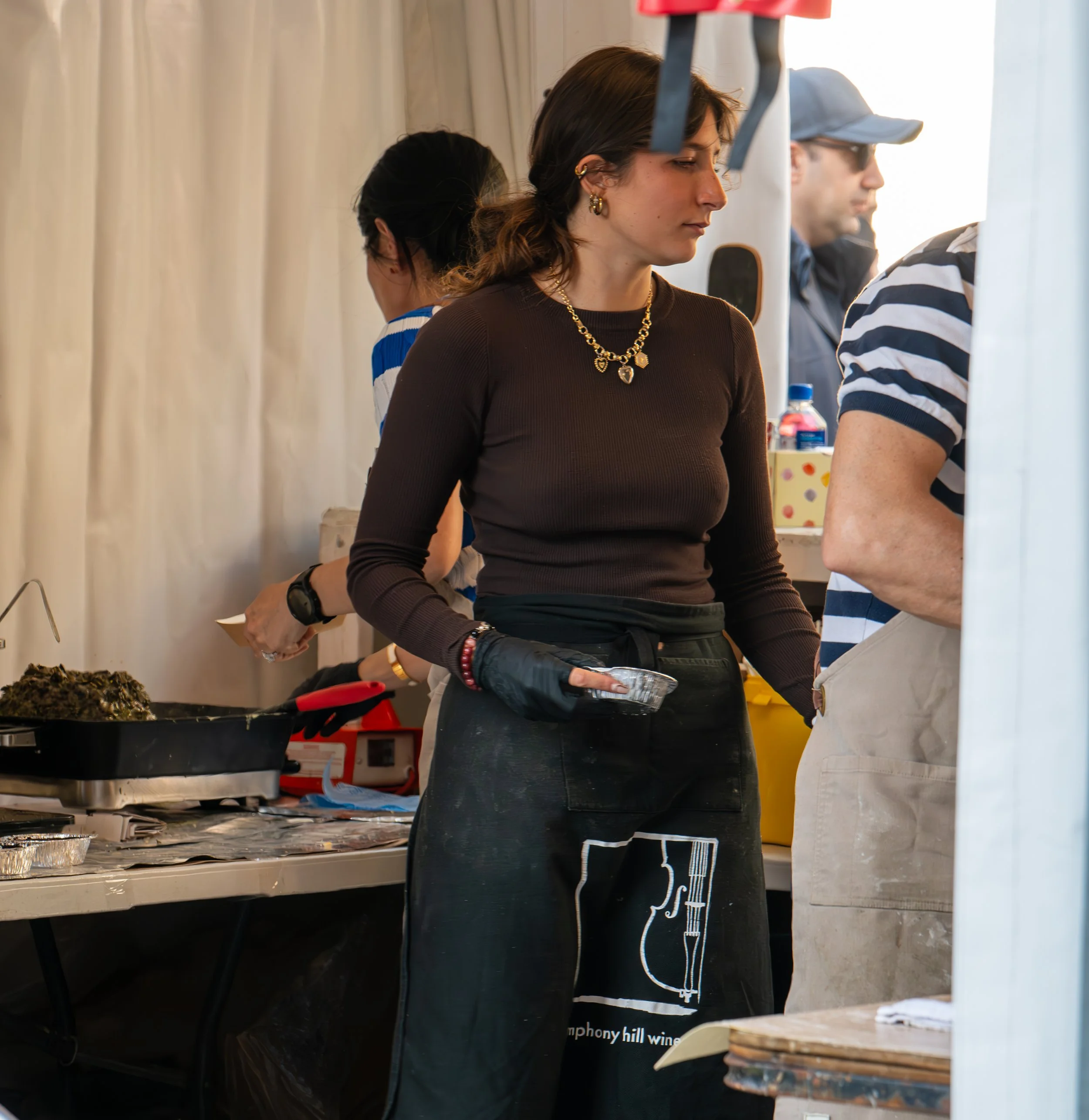 A woman with dark hair wearing a brown long-sleeve shirt, black gloves, and a black apron with white logo, standing in front of a counter with food, at a busy food stall or event.