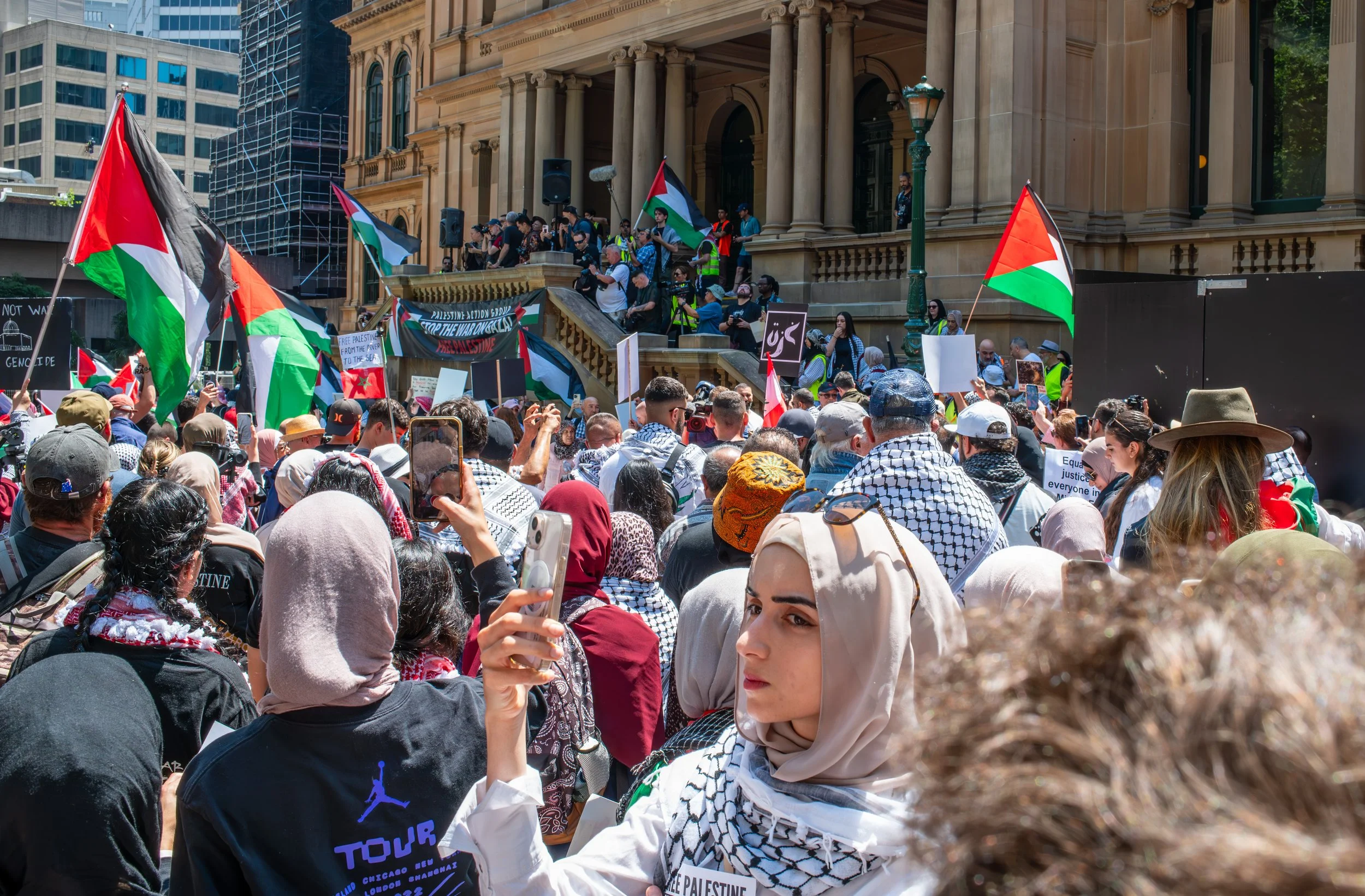 A large crowd of people protesting with Palestinian flags outside a historic building, some holding signs and taking photos.