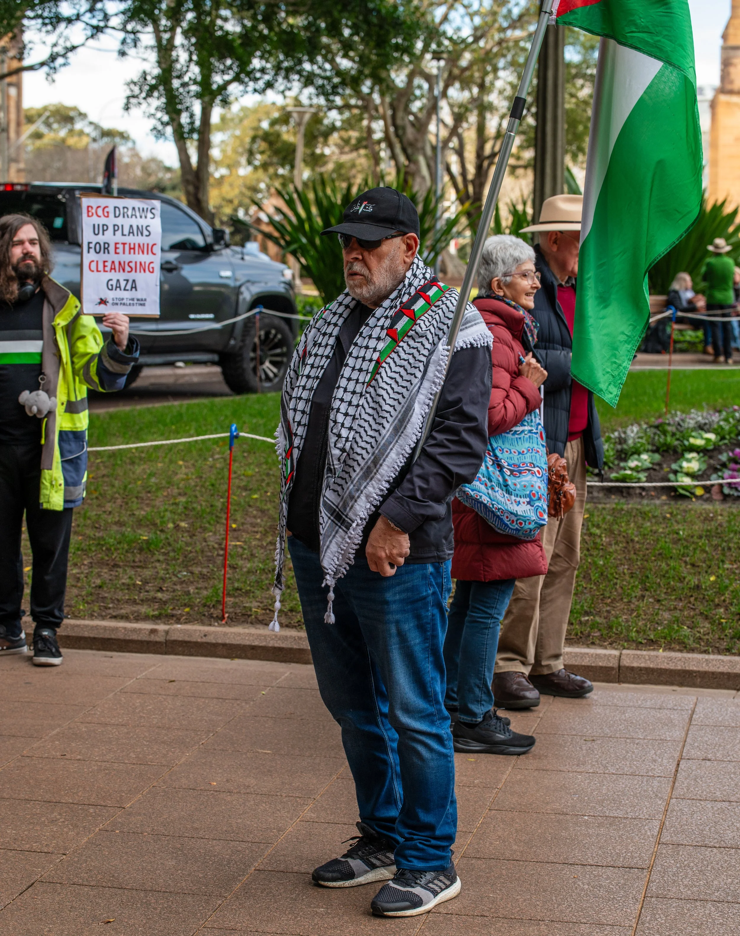 A protester holding a Palestinian flag stands with others in a park, with a sign in the background that reads 'BCG DRAW UP PLANS FOR ETHNIC CLEANSING GAZA'.