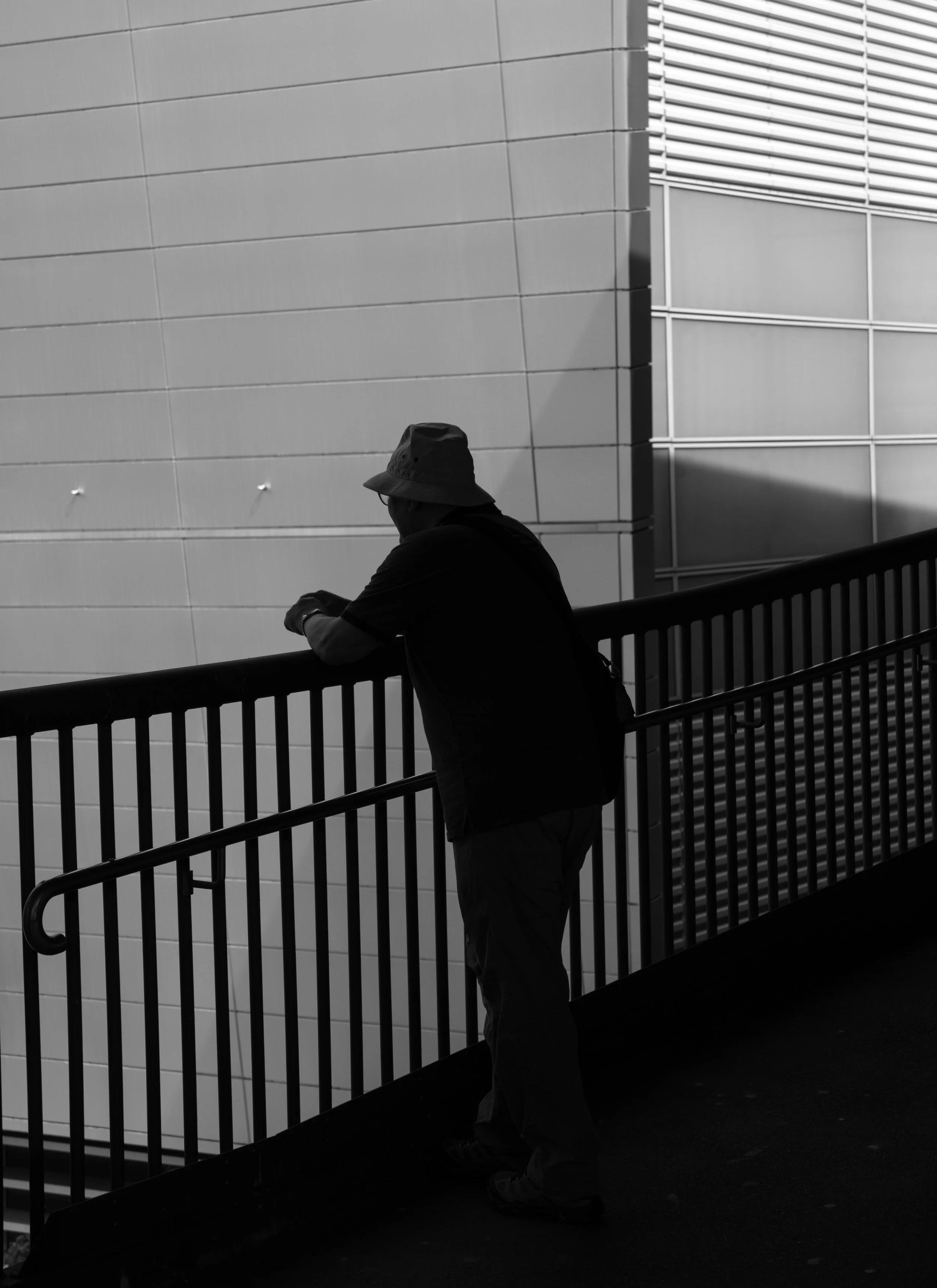 A silhouette of a man leaning on a railing, wearing a hat, against a modern building with varying textures and shadows.