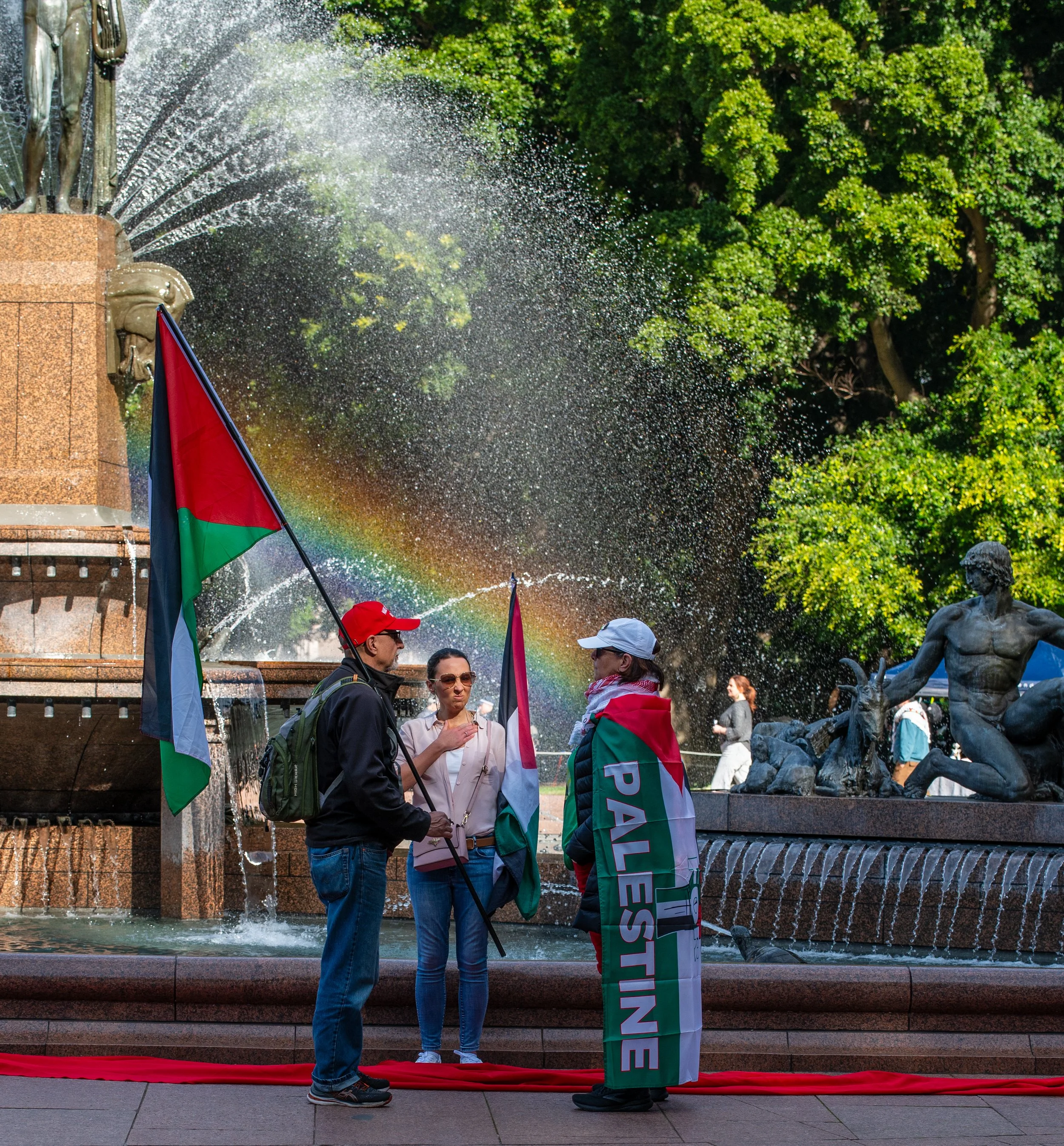 Three people standing by a fountain holding Palestinian flags and a Palestinian flag draped over one person's shoulders, with a rainbow appearing in the spray of the fountain, in a park with green trees in the background.