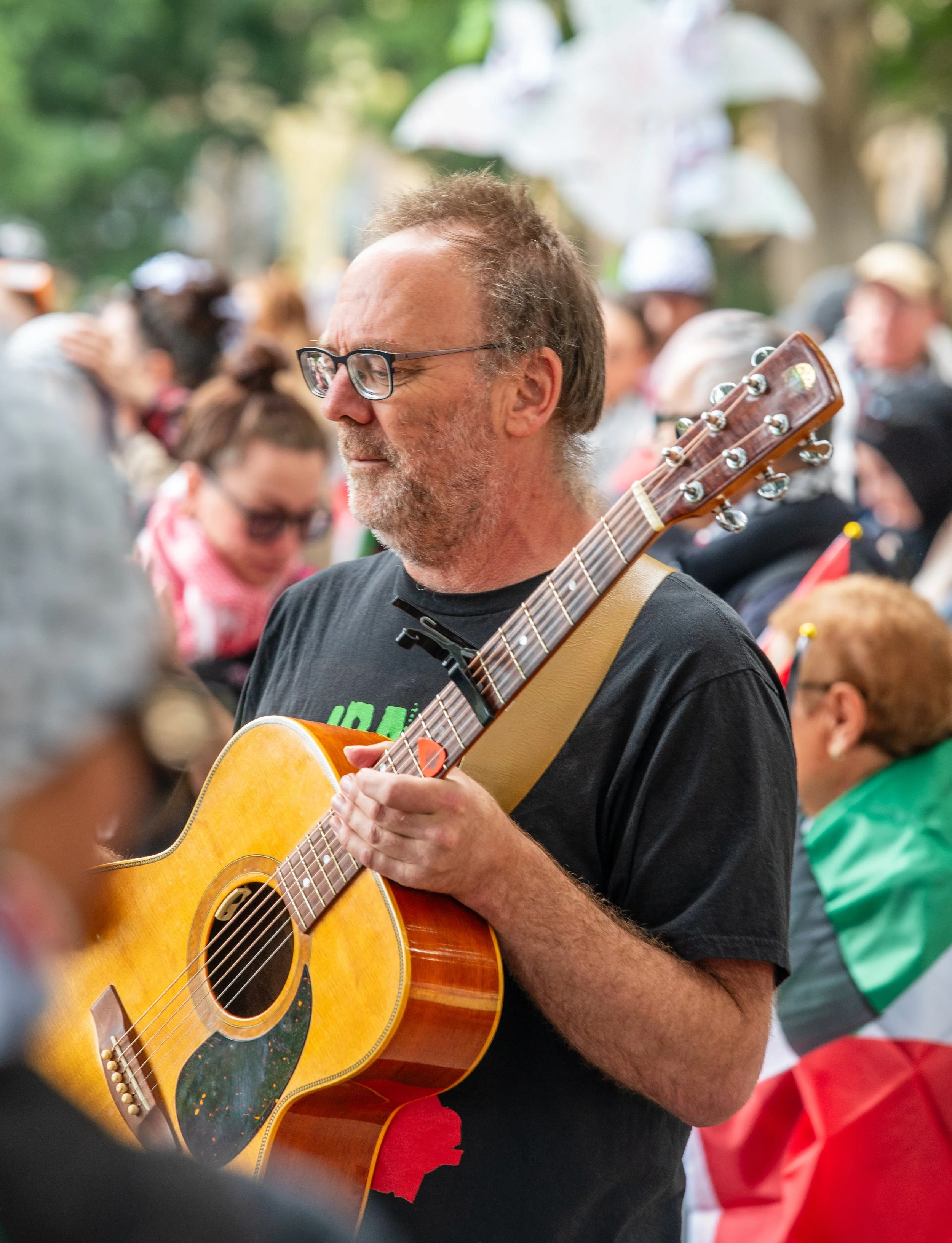 A man with glasses and a beard playing an acoustic guitar at an outdoor gathering, with a crowd of people in the background.