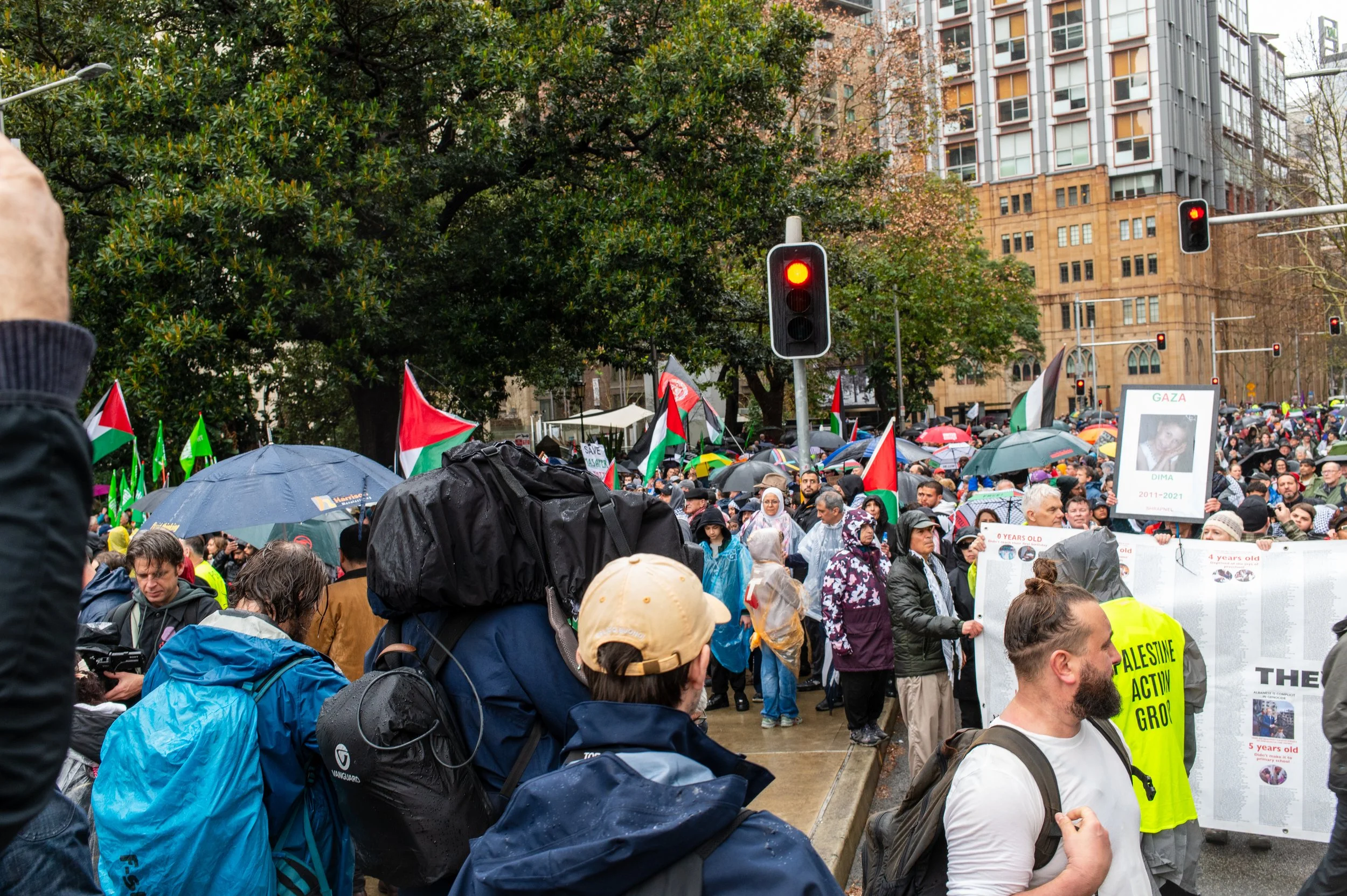 Crowd of people protesting outdoors with Palestinian flags and banners in the rain, some holding umbrellas, in an urban setting with tall buildings.