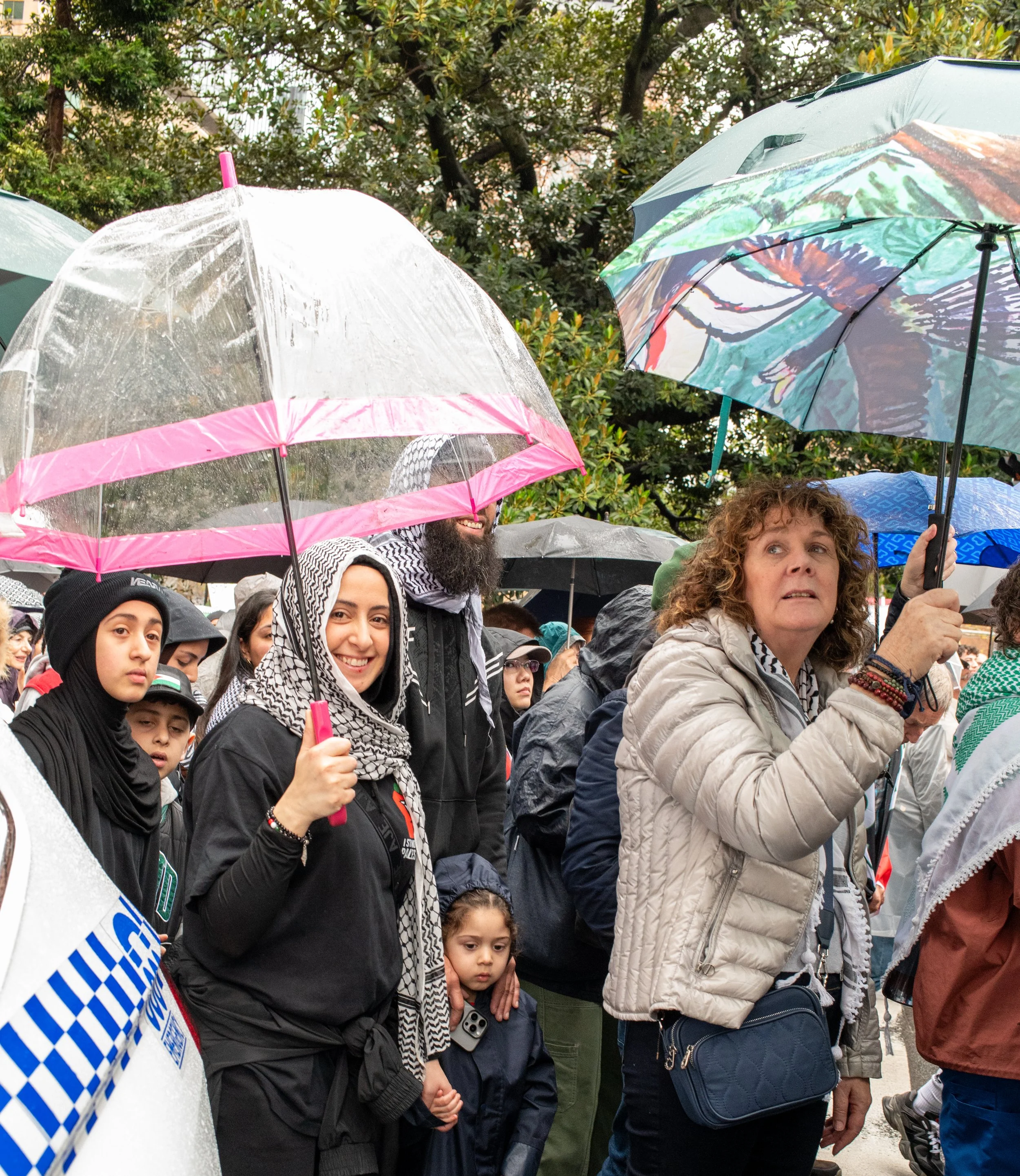 A crowd of people gathered outdoors holding umbrellas in the rain during a protest or rally, with diverse ages and backgrounds visible.