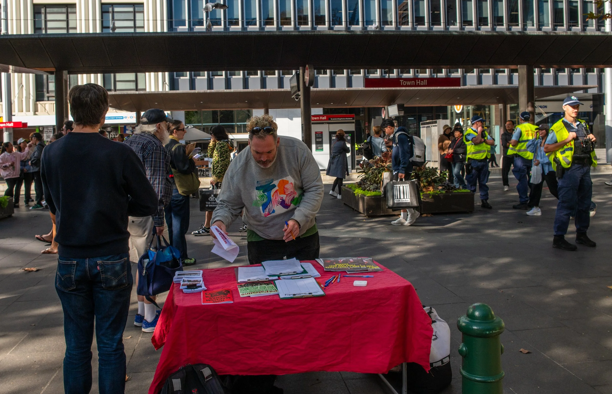 A busy outdoor scene at a city square with a table covered in red cloth setting up pamphlets, surrounded by people, including several police officers, in front of a building with signs for Town Hall and various shops.