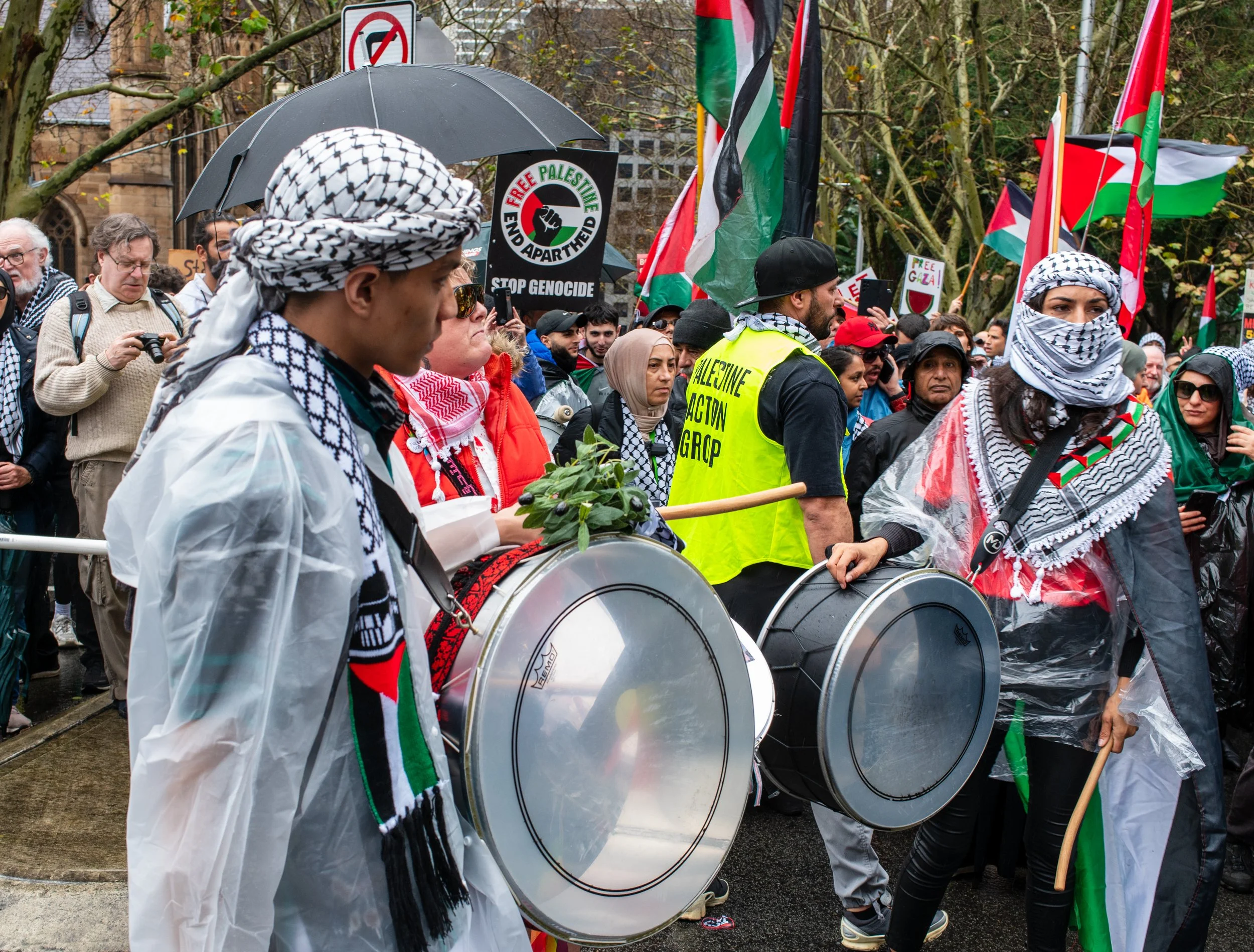 Pro-Palestinian protesters with flags and drums demonstrating outdoors, some wearing keffiyeh scarves and rain ponchos, holding signs saying 'Free Palestine' and 'Stop Genocide'.