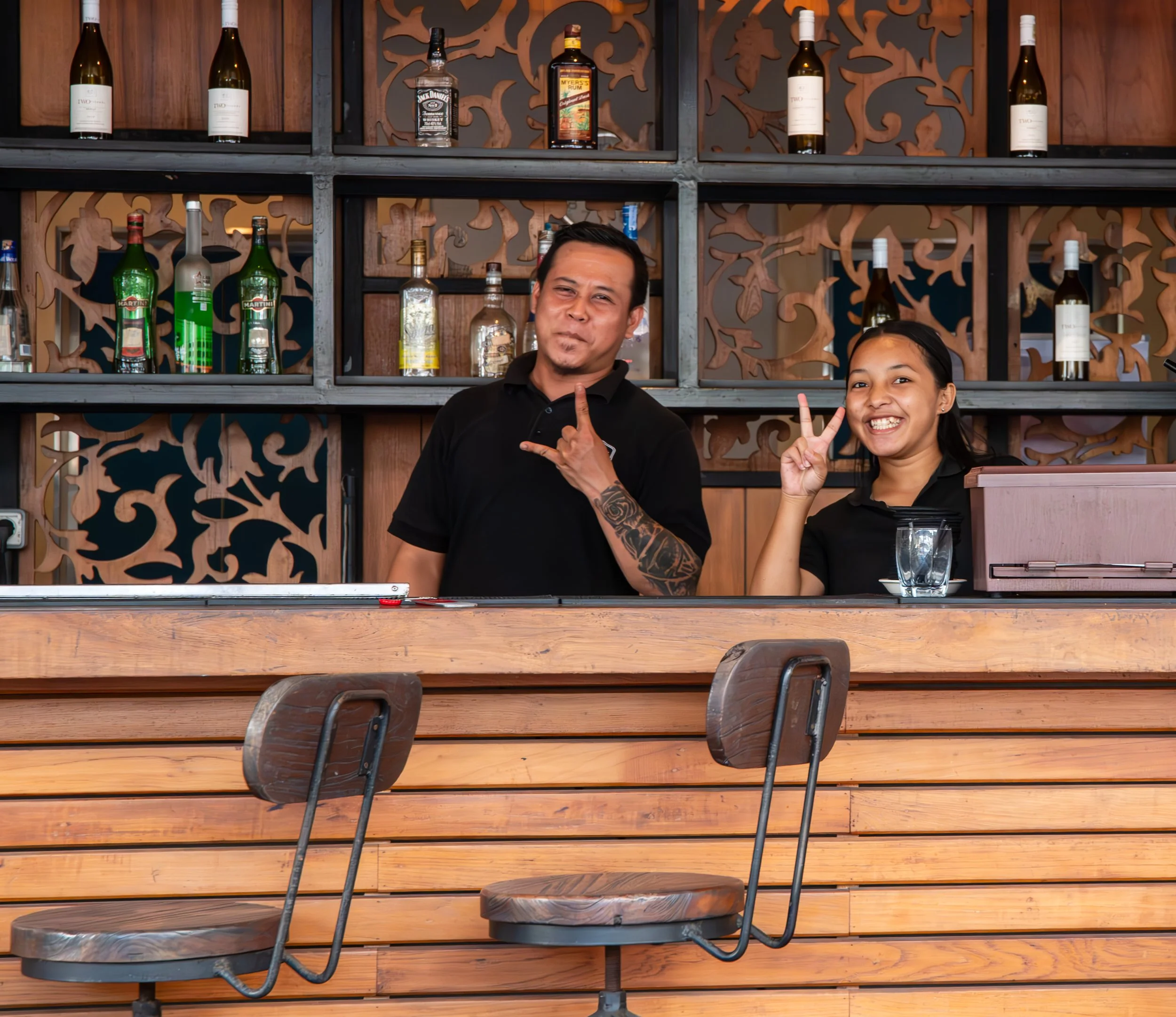 Two people, a man and a woman, smiling and making gestures while standing in front of a decorative wooden shelf with bottles of alcohol behind them. The man is making a rock sign with his right hand and the woman is showing a peace sign with her right hand. The setting appears to be a bar or restaurant.