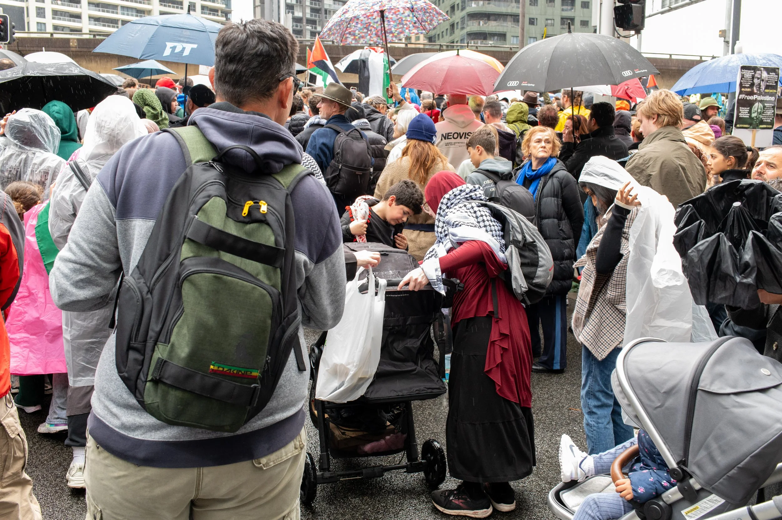 Crowd of people gathered at a protest or rally, many holding umbrellas in the rain, with diverse attire including raincoats and headscarves, and some carrying backpacks and using strollers.