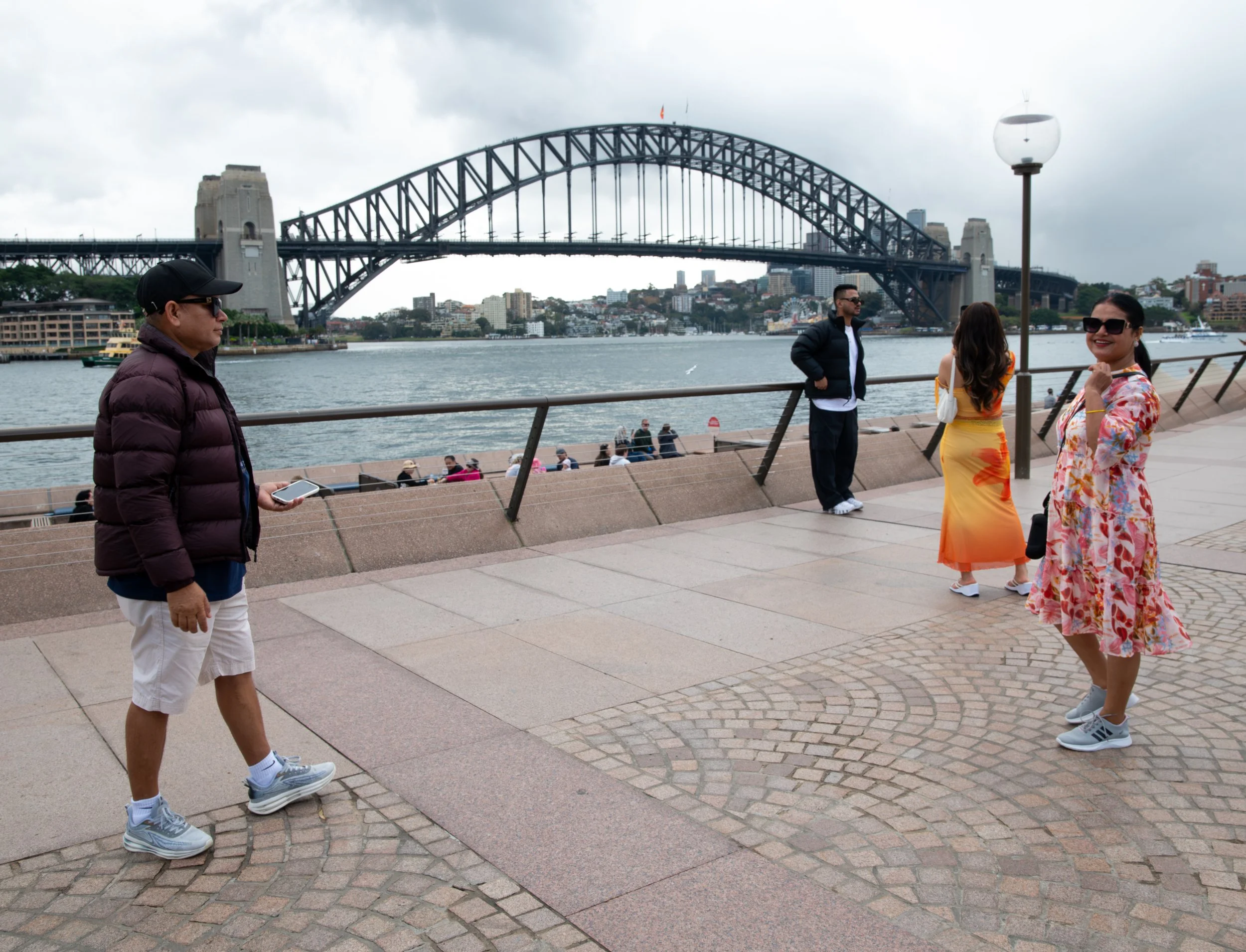 People walking and standing along a waterfront promenade with the Sydney Harbour Bridge in the background.
