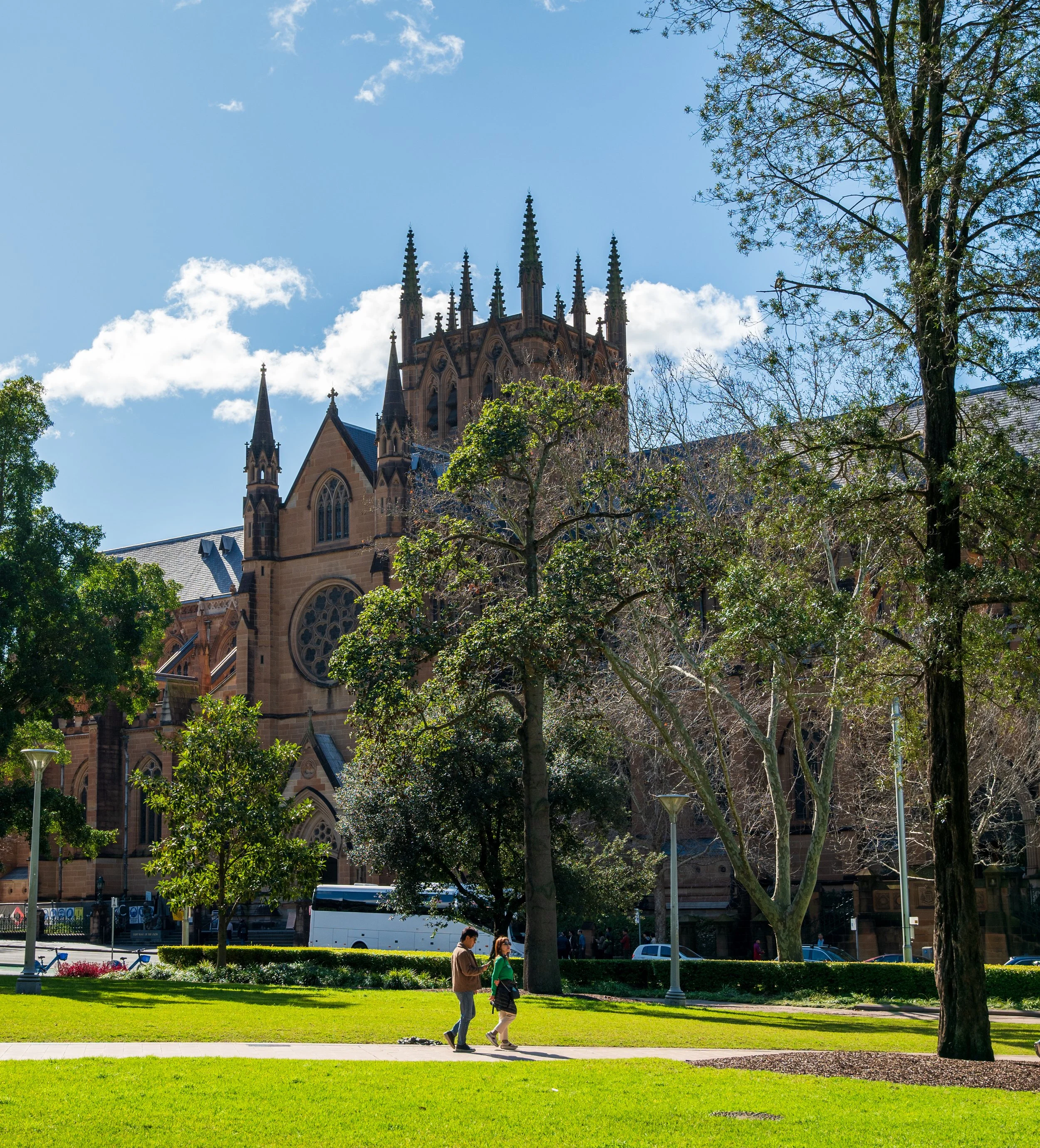 People walking on a grassy park with trees, in front of a large stone Gothic cathedral with spires, pointed arches, and rose windows, under a bright blue sky with clouds.