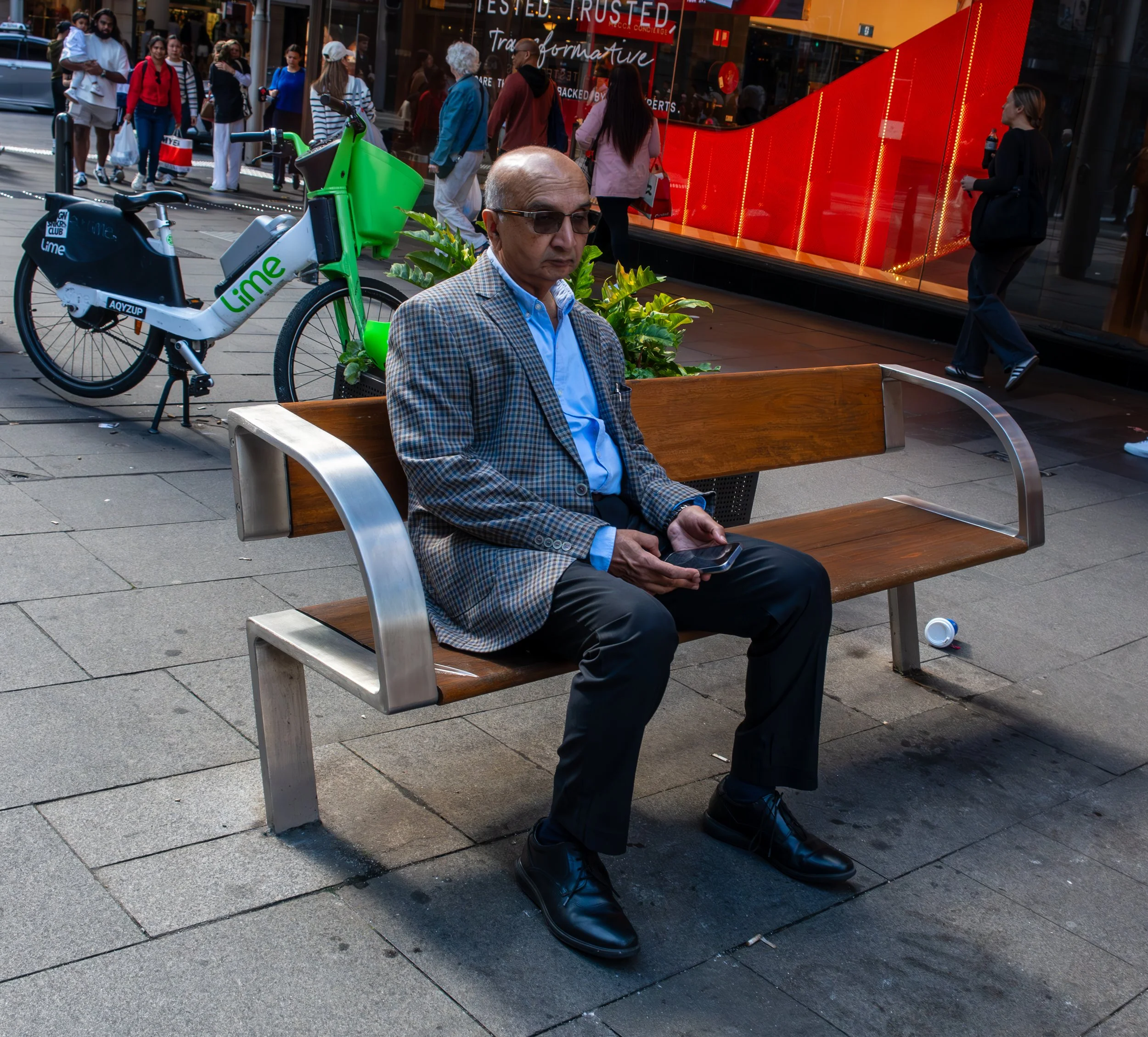 A man with glasses and a checkered blazer sits on a bench on a city sidewalk, holding a smartphone. Behind him are pedestrians, a green bike with a Lime logo, and a bright red storefront display.