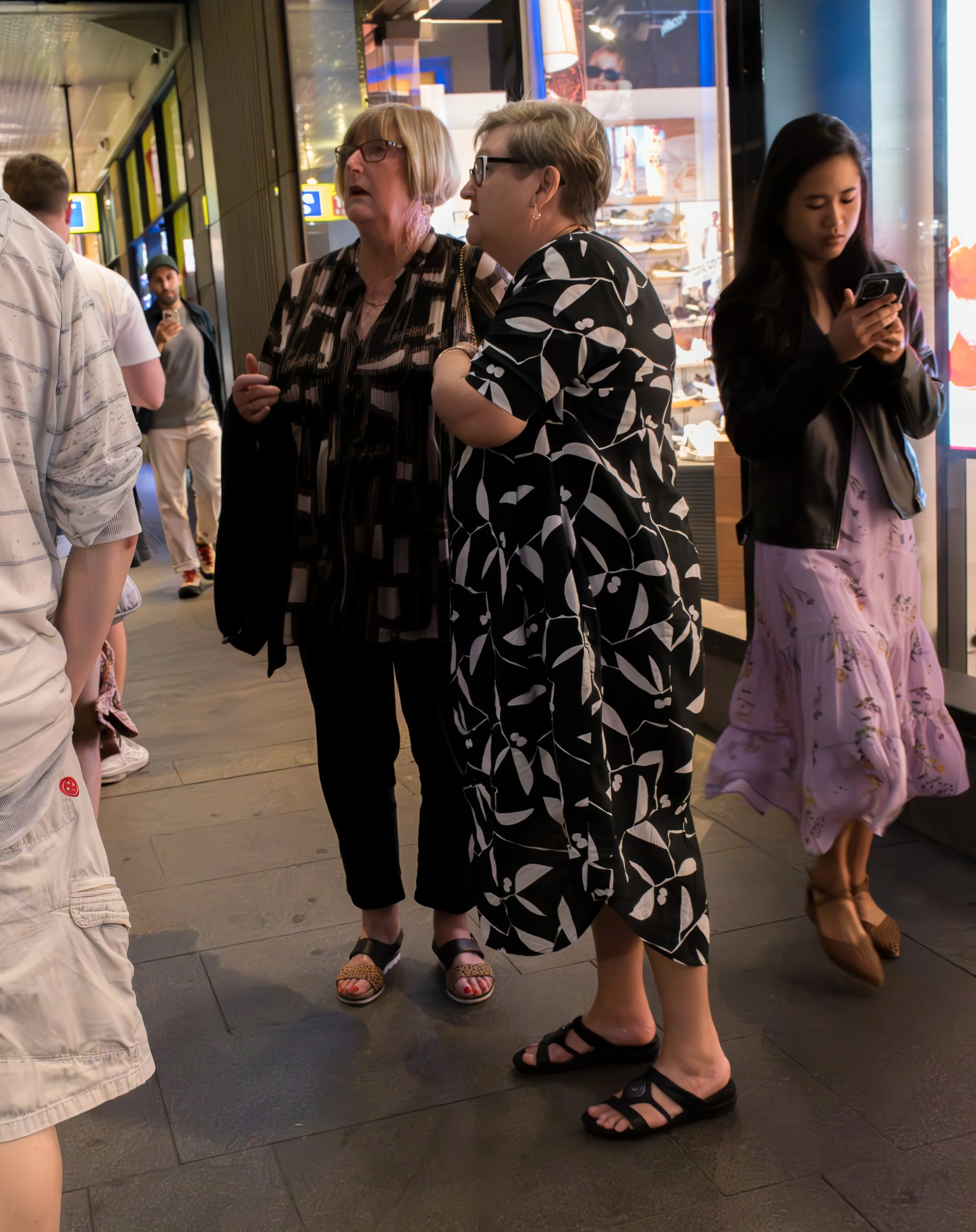 Two women having a conversation on a city sidewalk at night, surrounded by other pedestrians, with a shop window in the background.