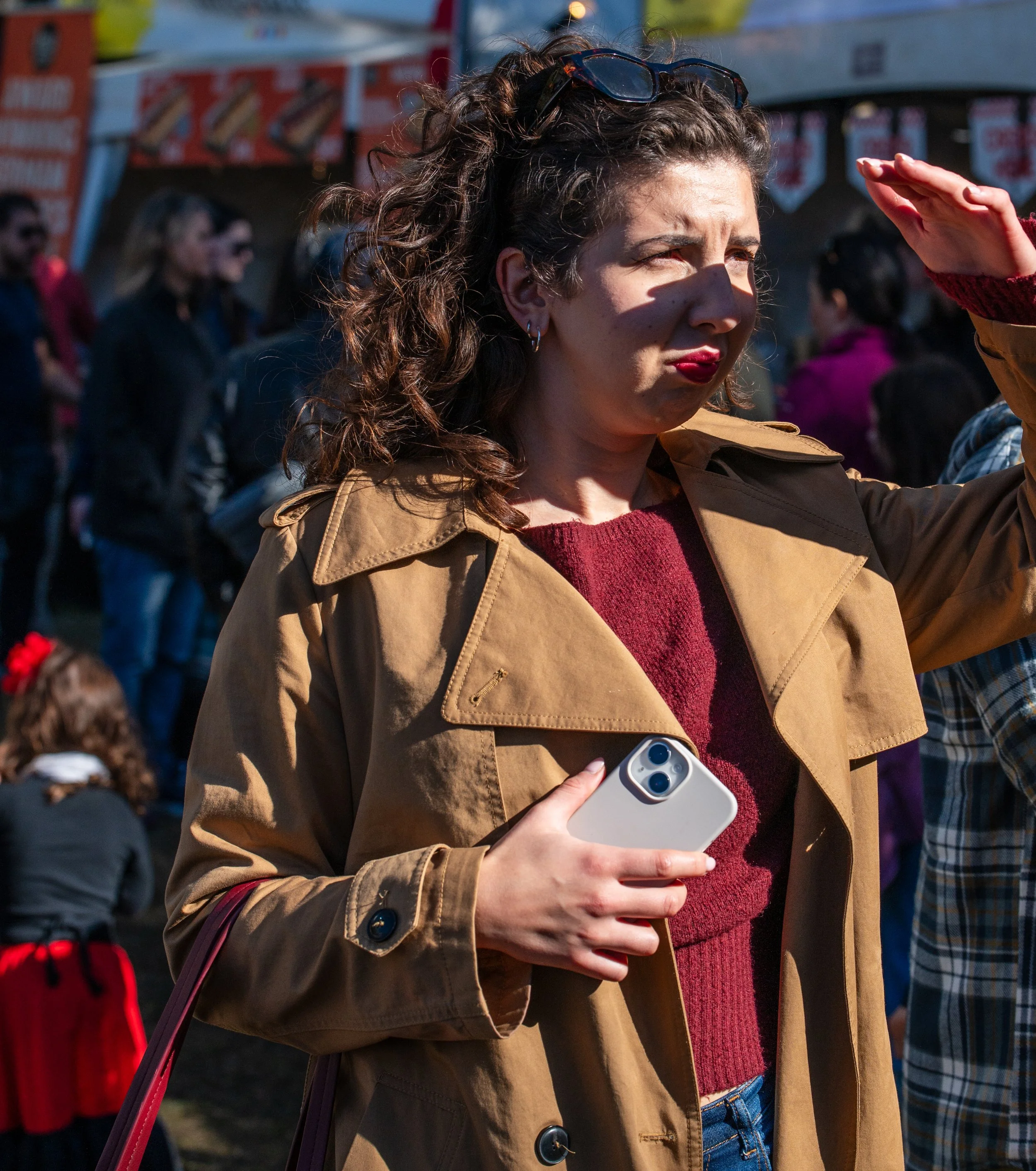 A woman with curly brown hair, wearing a tan trench coat and red sweater, is holding a smartphone and shielding her eyes from the sun at an outdoor event with crowd and food stalls.