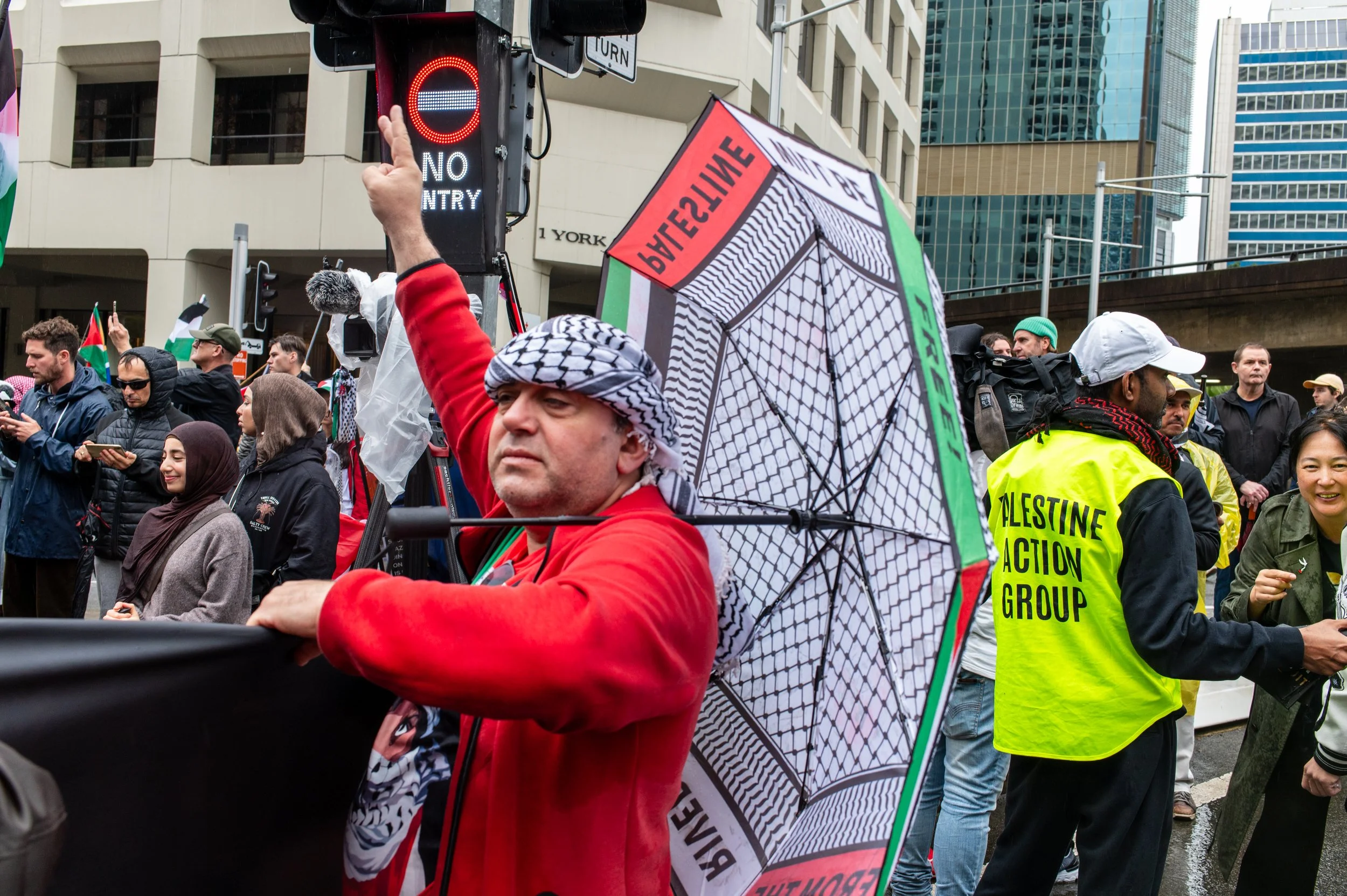 A protest scene with people holding signs and flags in an urban area. A man in the foreground is wearing a red jacket and a keffiyeh, holding a large rainbow-colored umbrella with Palestinian flags and text. There are other protesters, some wearing yellow vests, some smiling, and a