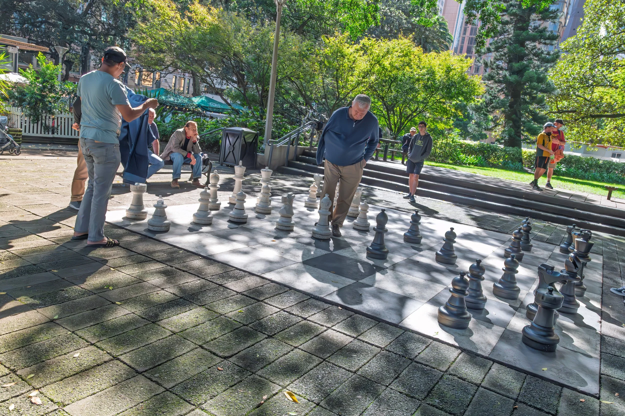 People playing chess outdoors in a park on a sunny day, with trees and buildings in the background.