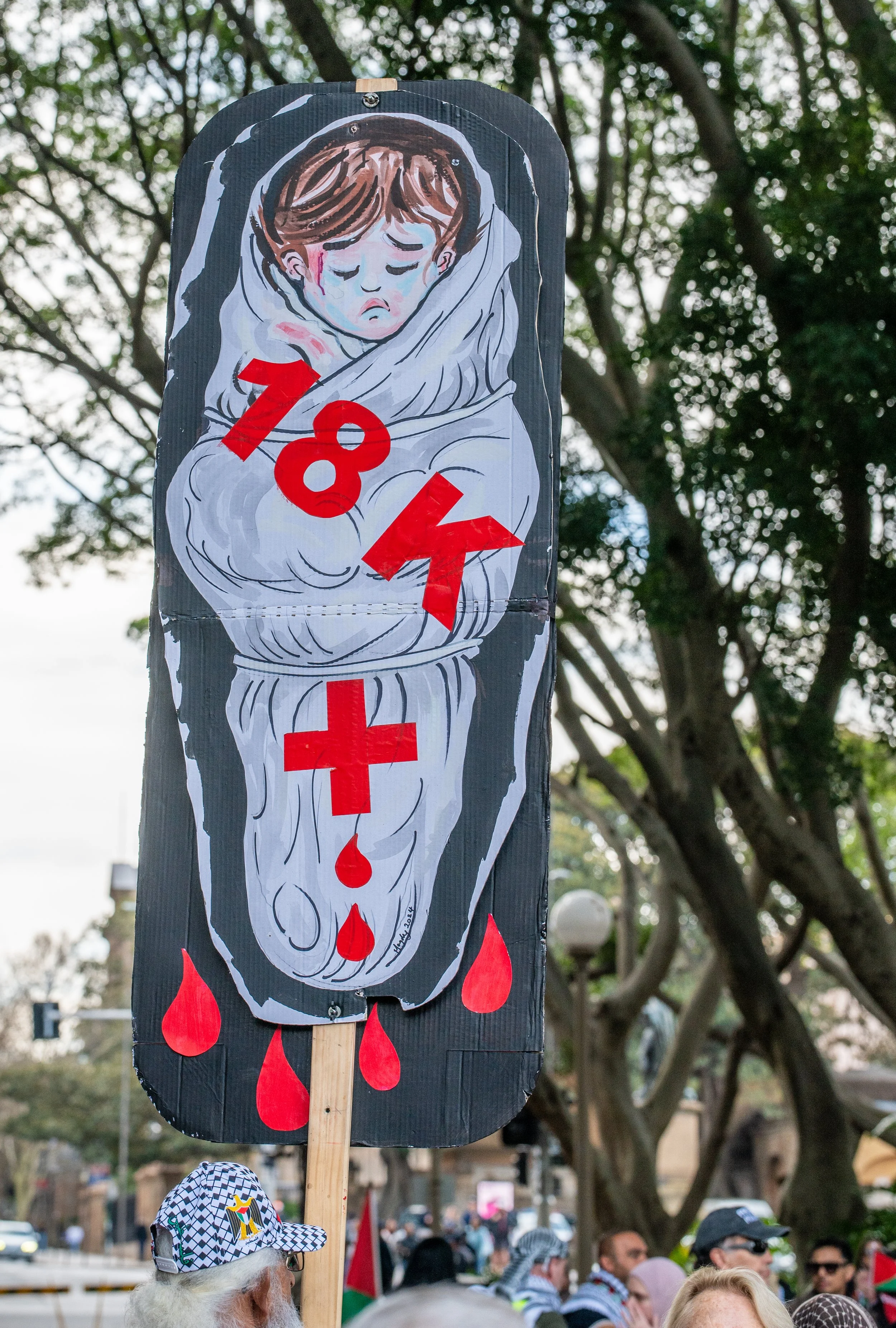 A protest sign depicting a sad, imprisoned child wrapped in bandages, with red warning symbols and drops of blood, held by a person in a crowd.