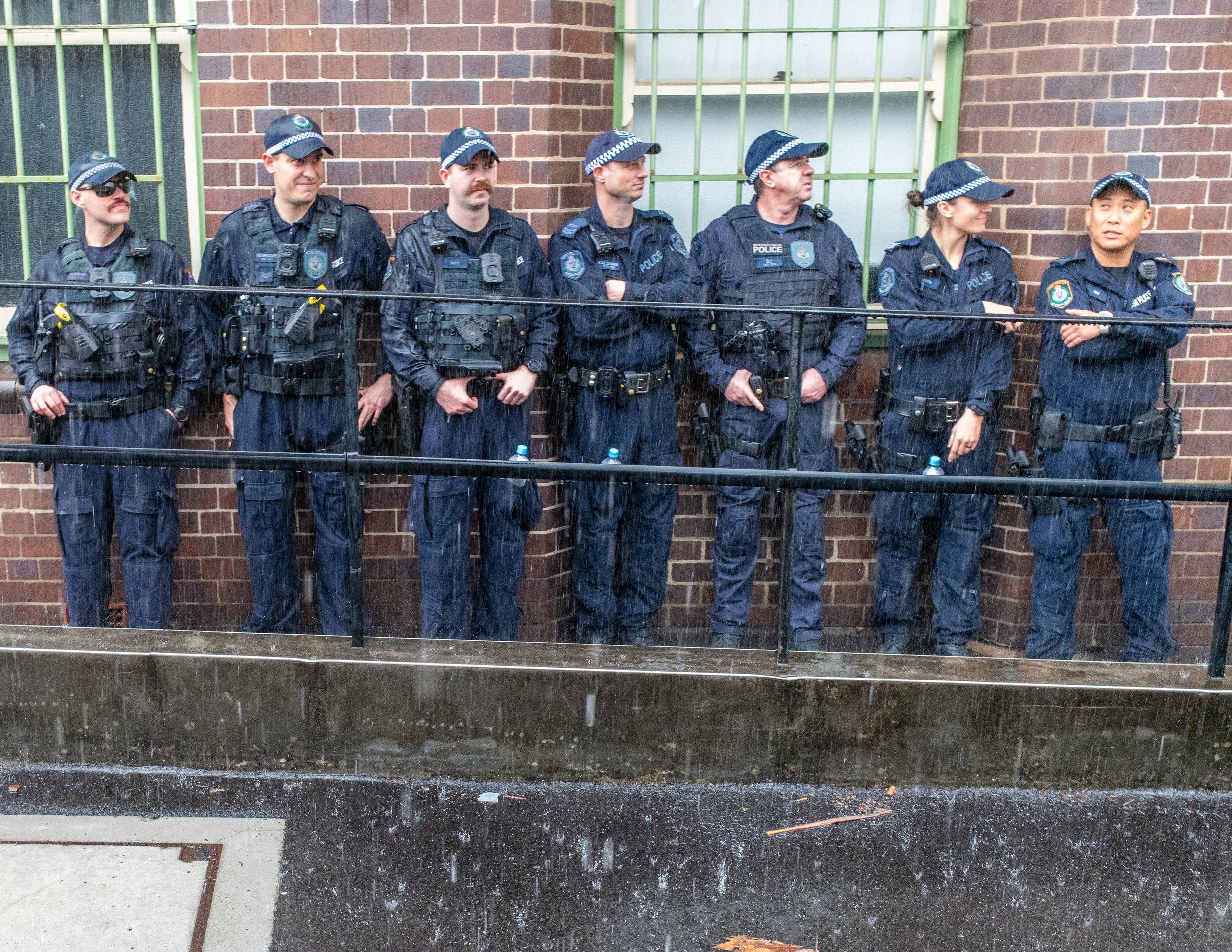 Line of police officers standing in the rain against a brick wall.
