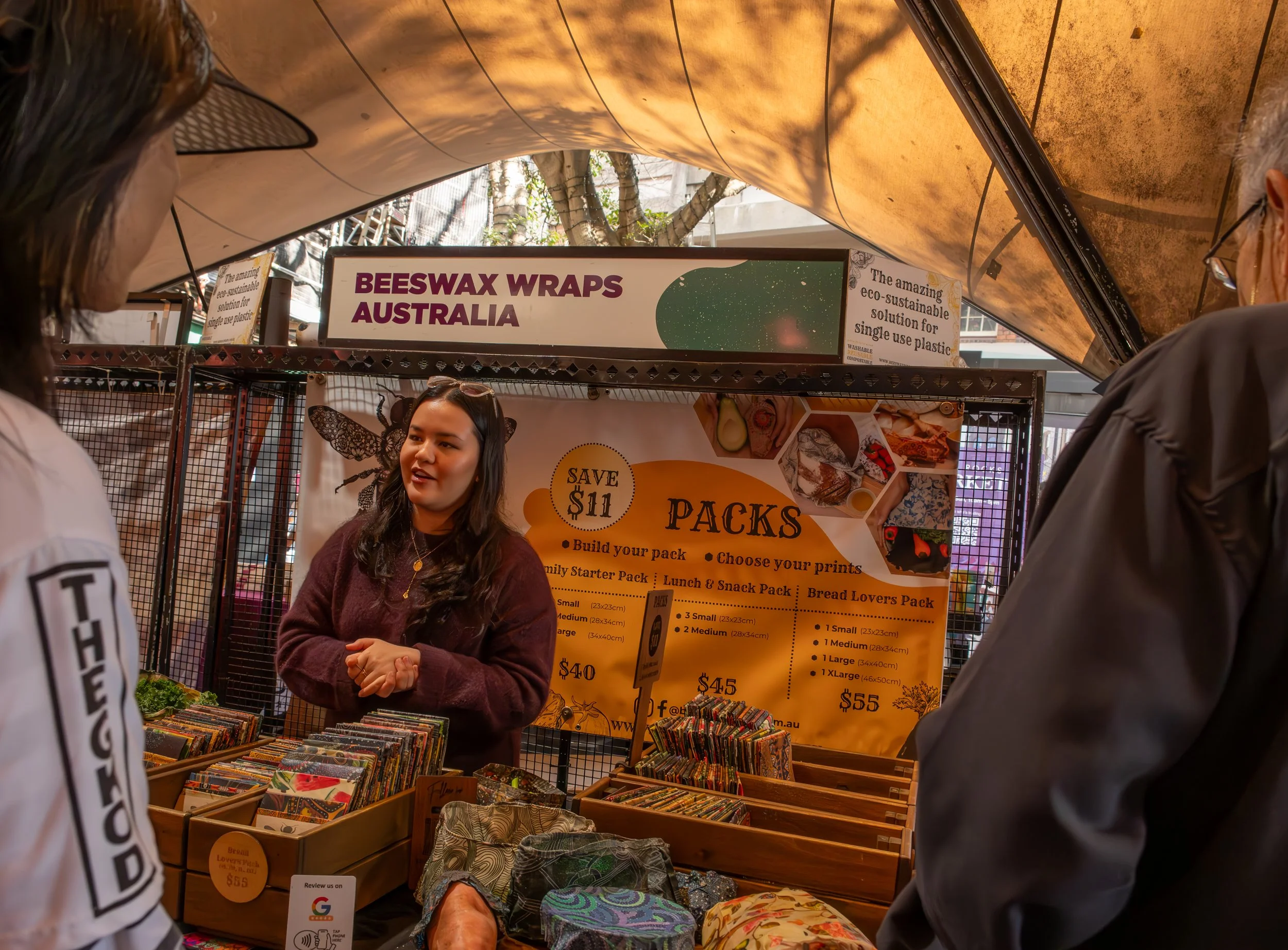 A woman at a stall selling eco-friendly beeswax wraps in Australia, with two customers in front. The stall displays colorful patterned wraps and has a sign promoting eco-sustainable single-use plastic solutions.