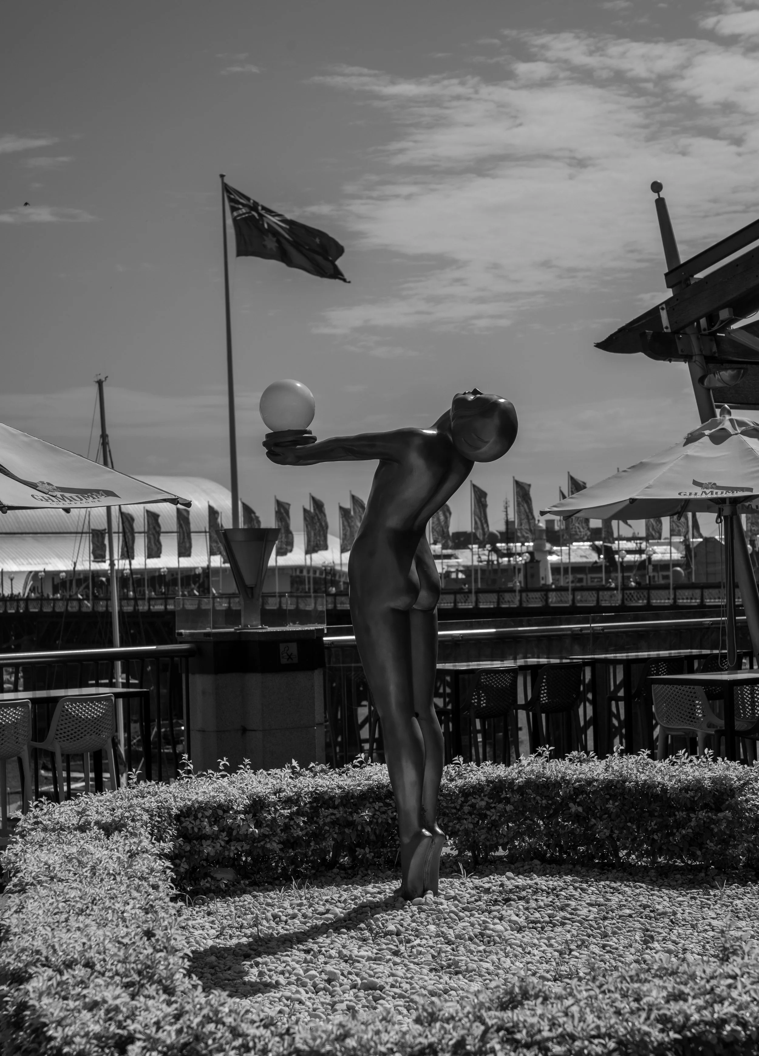 A sculpture of a stylized human figure holding a sphere with a flag of Australia in the background, set outdoors with umbrellas and flags along a waterfront.