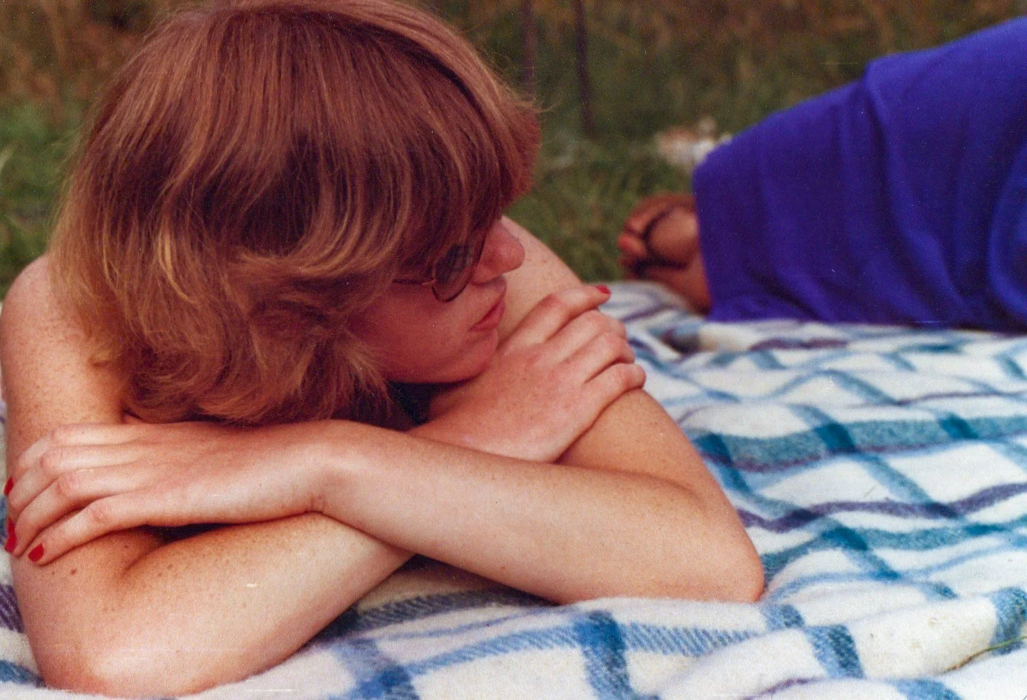 A woman with red hair and glasses lying on her side with her arms crossed, resting her head on her arms, on a striped blanket outdoors.