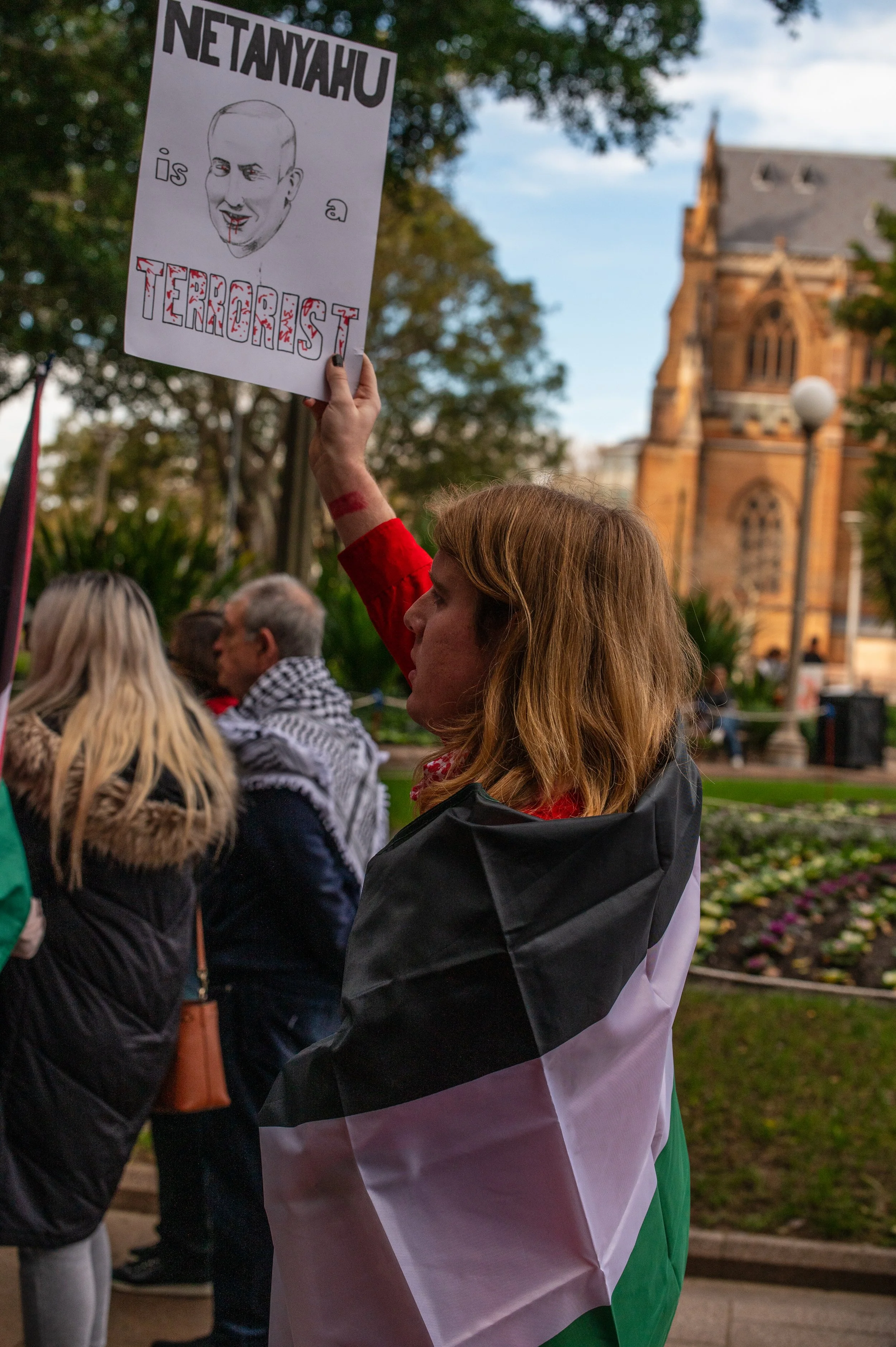 A woman at a protest holding a sign that depicts a caricature of Netanyahu with the words 'Netanyahu is a terrorist'. She is draped in a flag with black, white, and green colors, and there are other protesters and a historic building in the background.