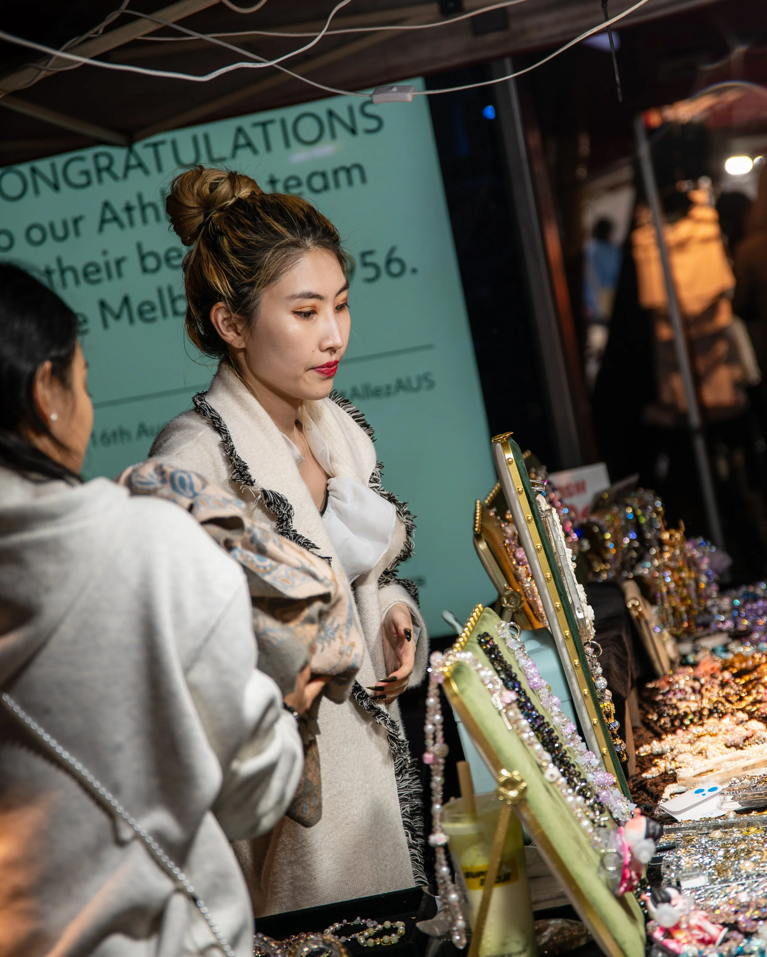Two women at an outdoor jewelry stall, browsing and inspecting various accessories and jewelry items displayed on tables and stands.