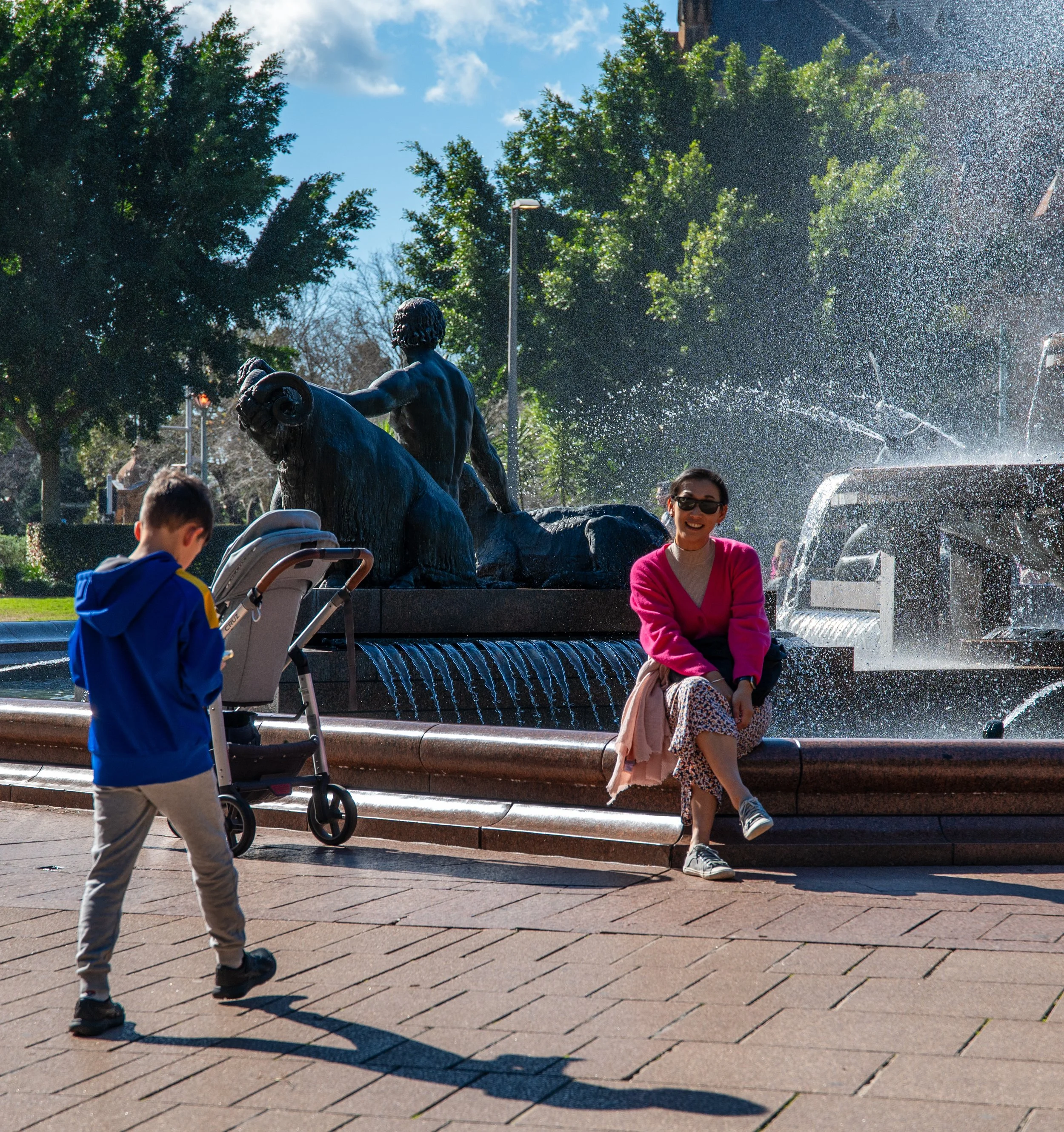 A woman sitting on the edge of a fountain with a sculpture, a young boy walking nearby with a stroller in a park setting with trees and a clear blue sky.