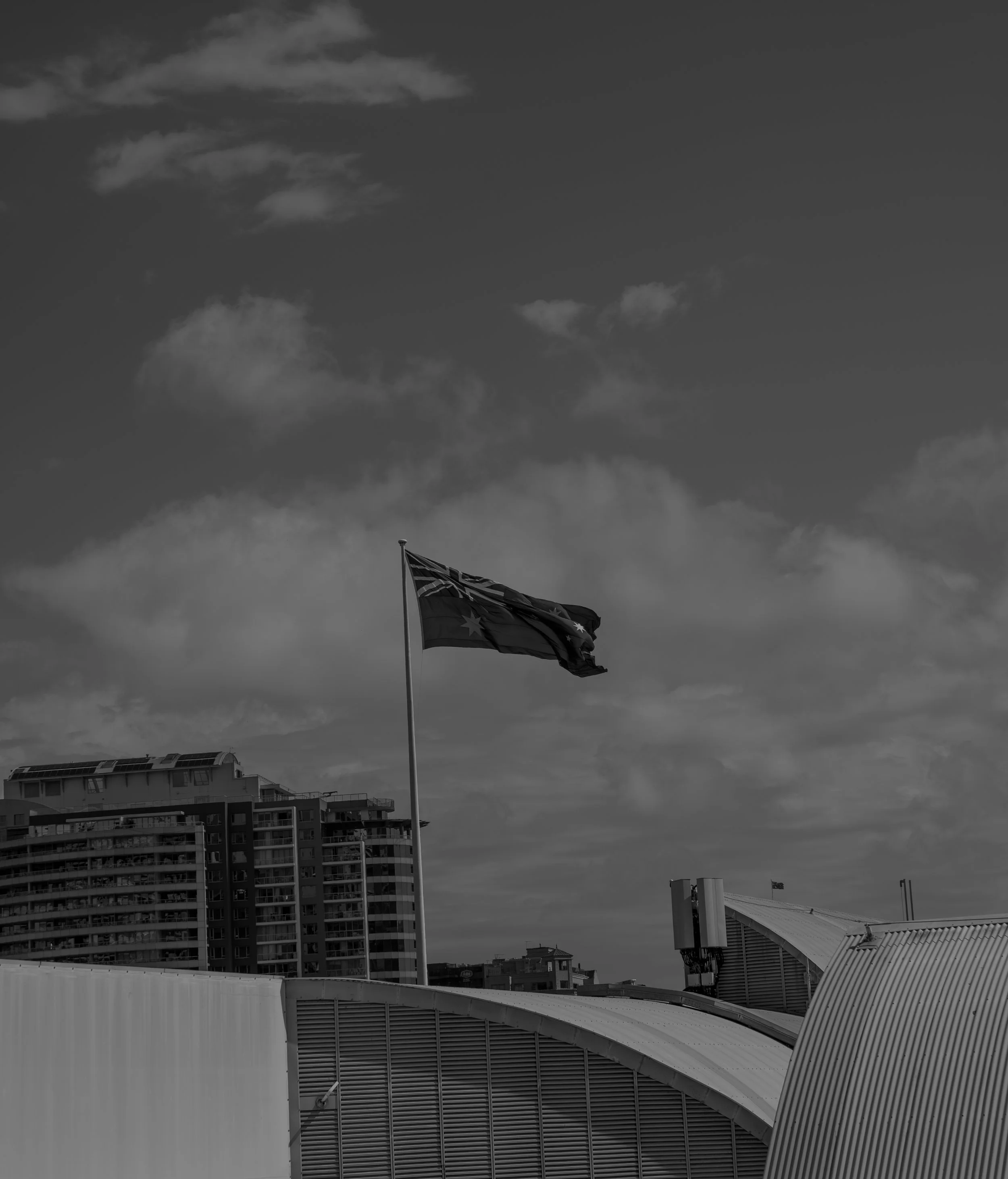 Black and white photo of a flag flying on a flagpole, with a background of modern buildings and a cloudy sky.