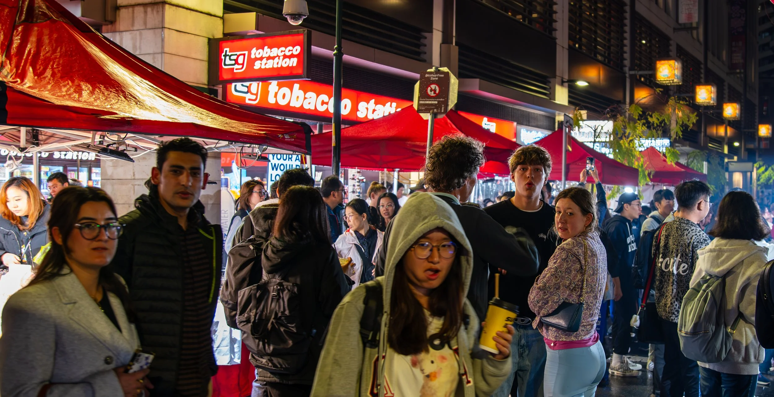 Crowded city street at night with people walking past a tobacco store and food vendors under red tents, illuminated signage, and lit street lamps.