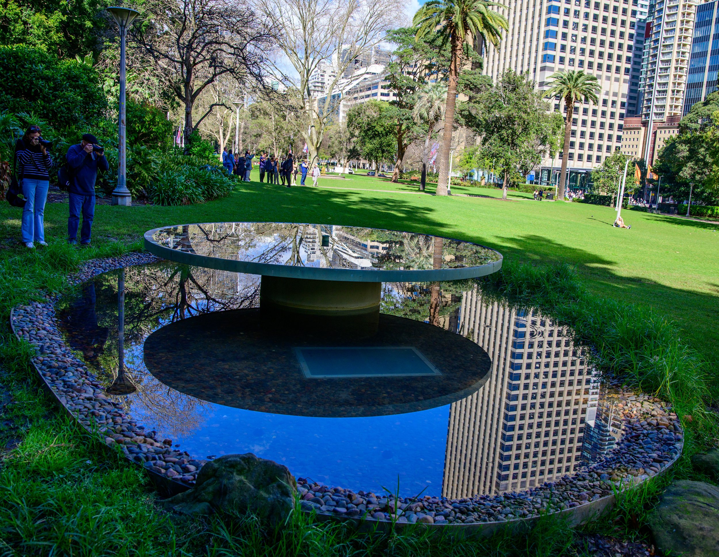 Public park with modern art installation reflecting tall buildings, surrounded by trees and people walking or sitting.