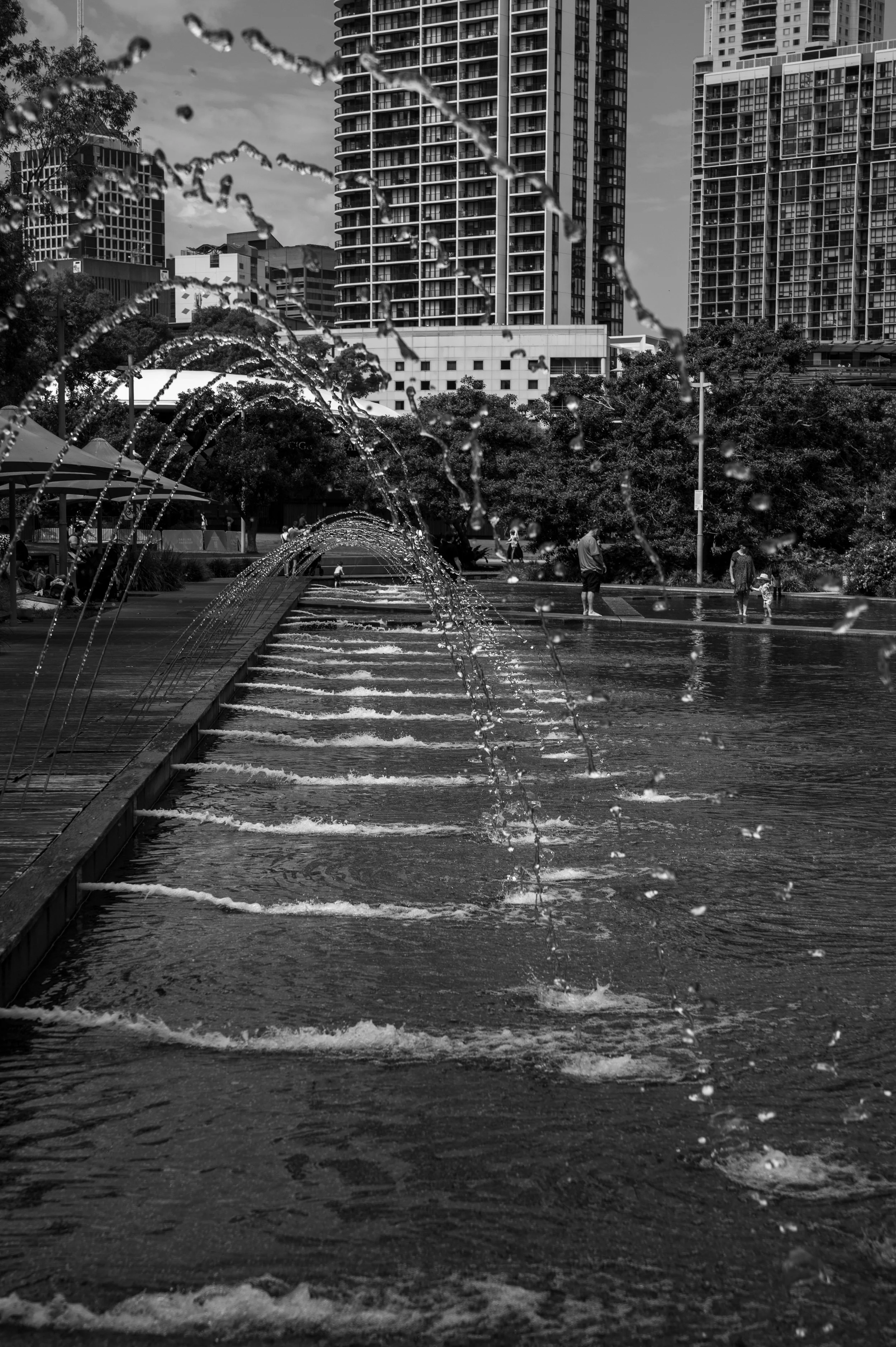 Black and white photo of a city park with fountains in the foreground and tall apartment buildings in the background.