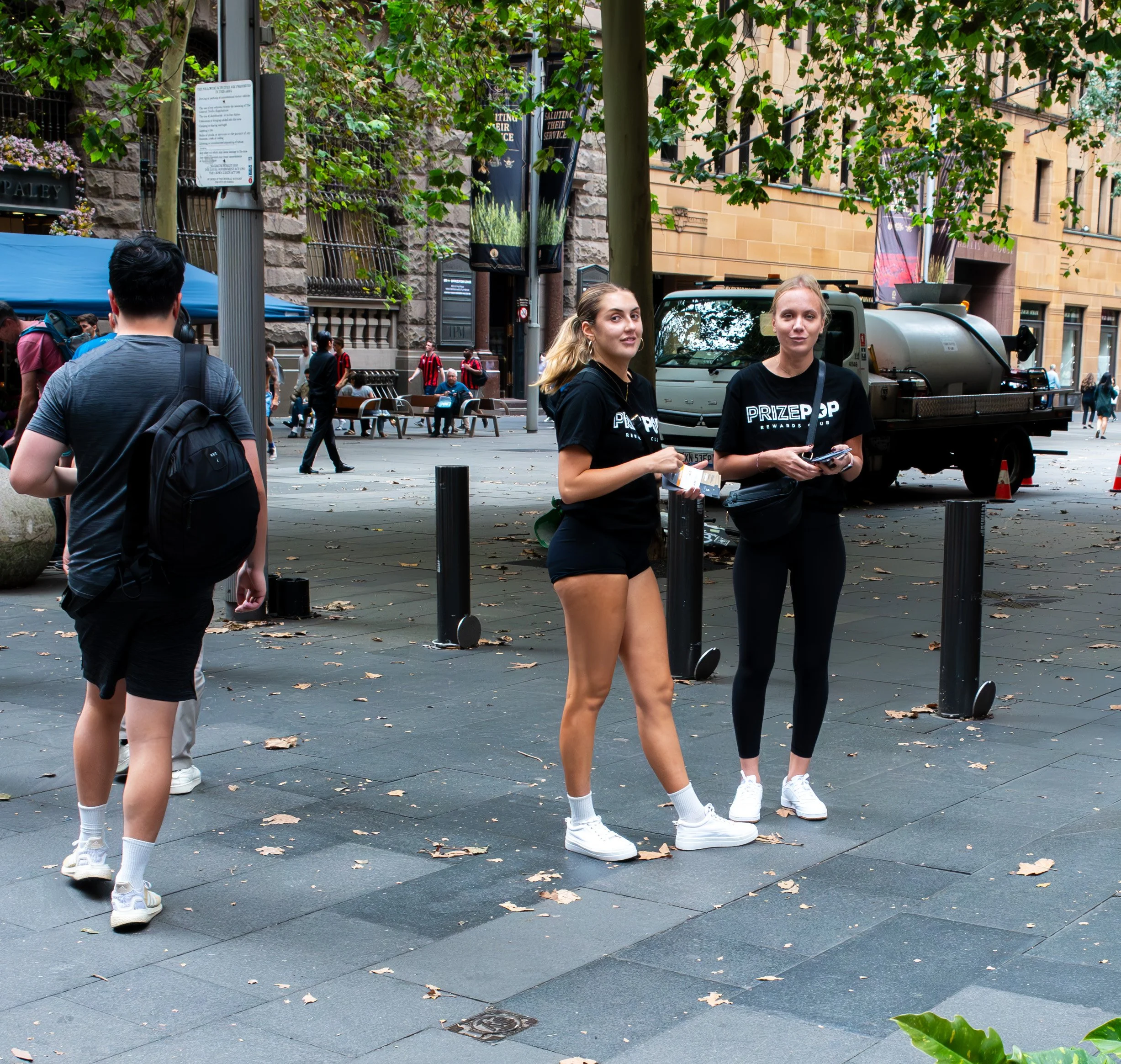 Two young women in Black PrizePop t-shirts standing on a city sidewalk, holding flyers and talking to a nearby man with a backpack. Other pedestrians and a water truck are visible in the background.