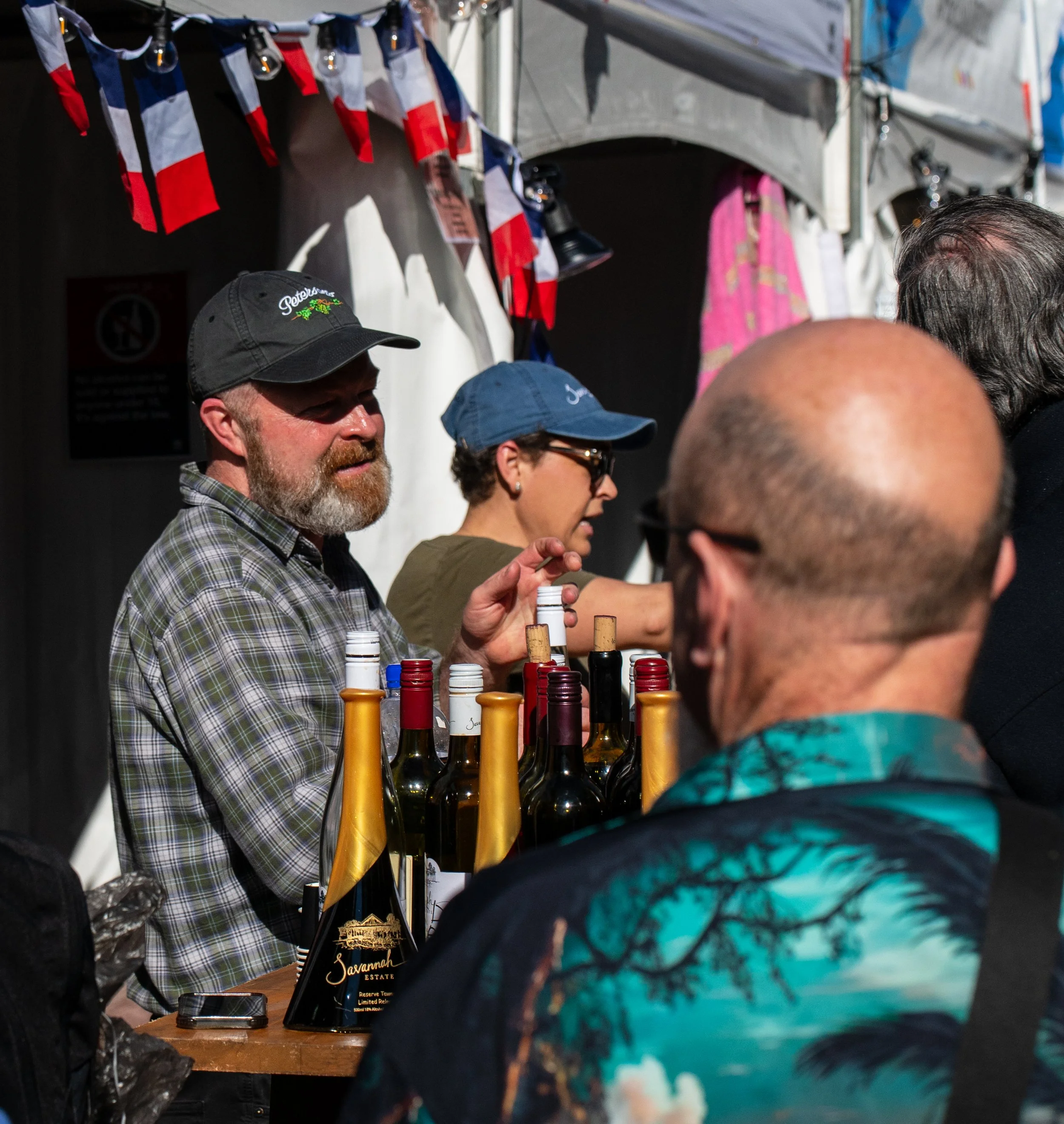 People at a wine booth at an outdoor event, with bottles of wine on the table and small flags hanging above.