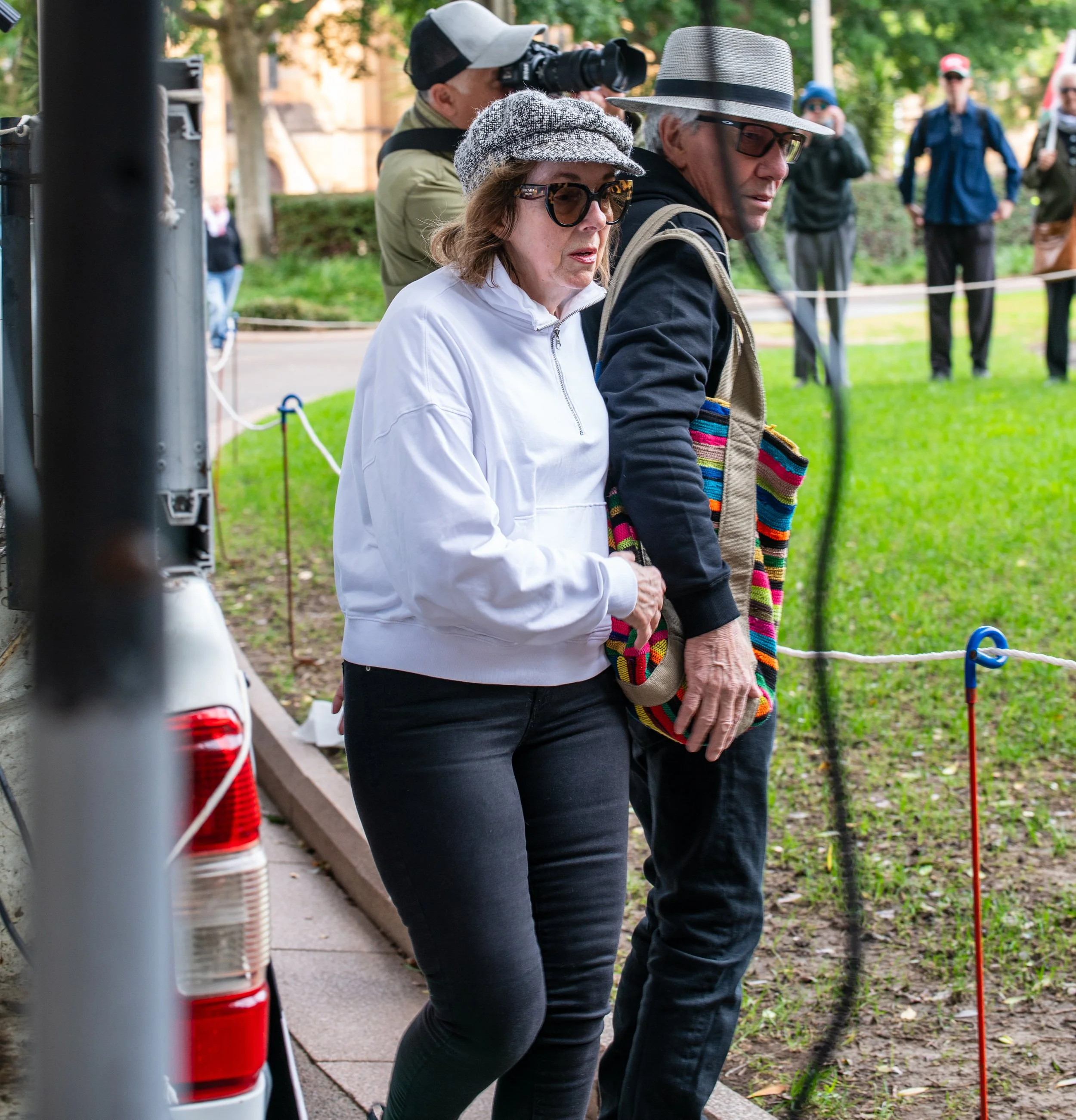 A group of people, including a woman and a man, are standing outdoors in a park-like setting. The woman is wearing sunglasses, a gray cap, and a white jacket. The man is wearing glasses, a gray fedora, a black jacket, and has a colorful bag over his shoulder. They appear to be observing something, with some people in the background also looking on.