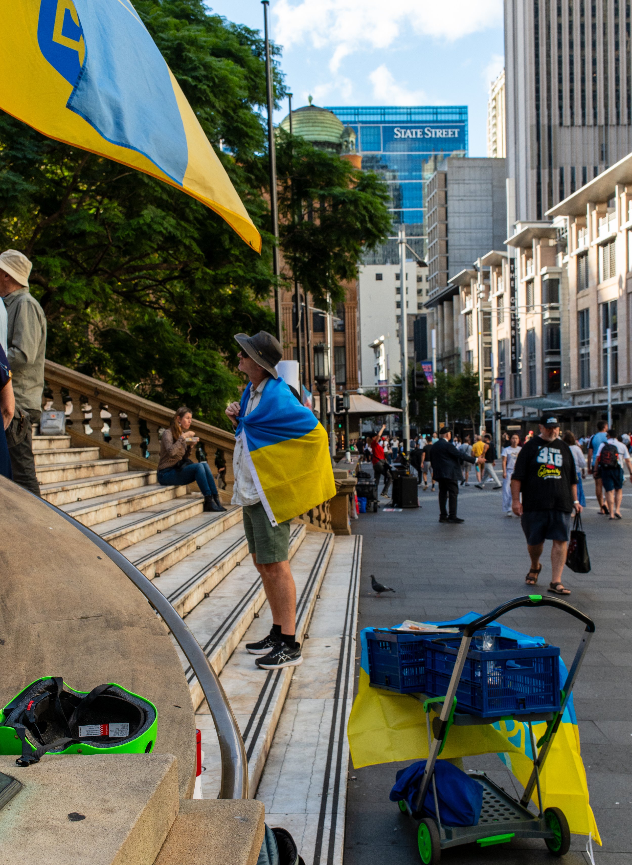 A man standing on stairs wrapped in a Ukrainian flag, in a busy city street with tall buildings and people walking.