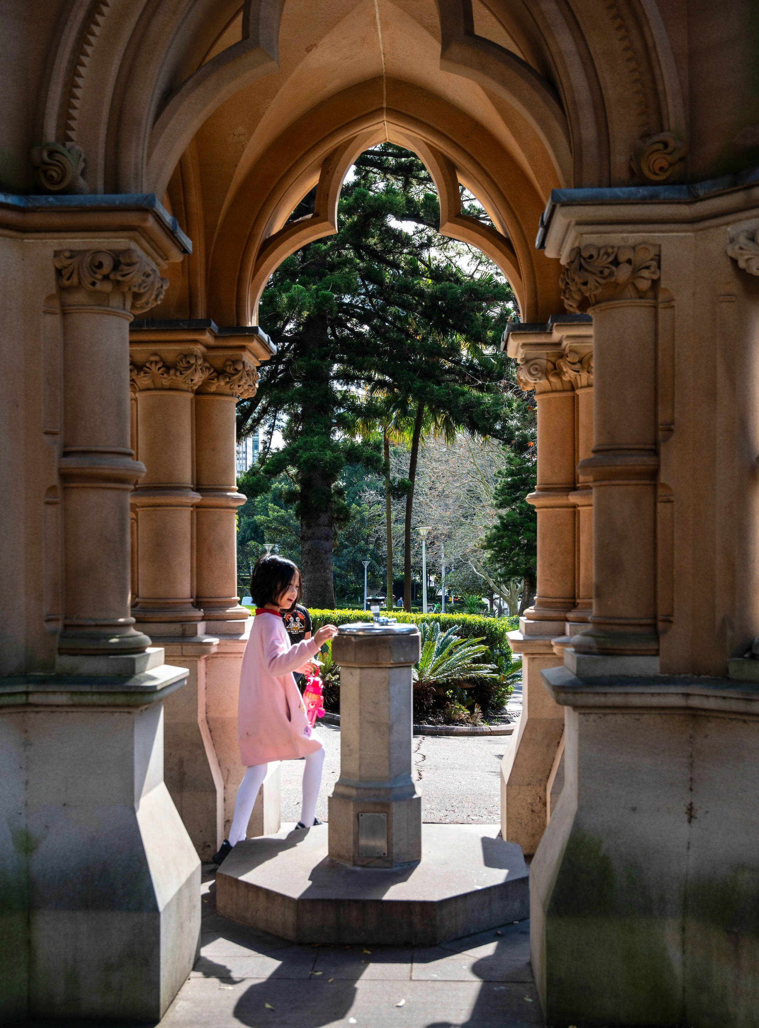 A young girl in a pink dress and white tights standing by a stone fountain under a decorative stone archway in a park with trees and greenery in the background.