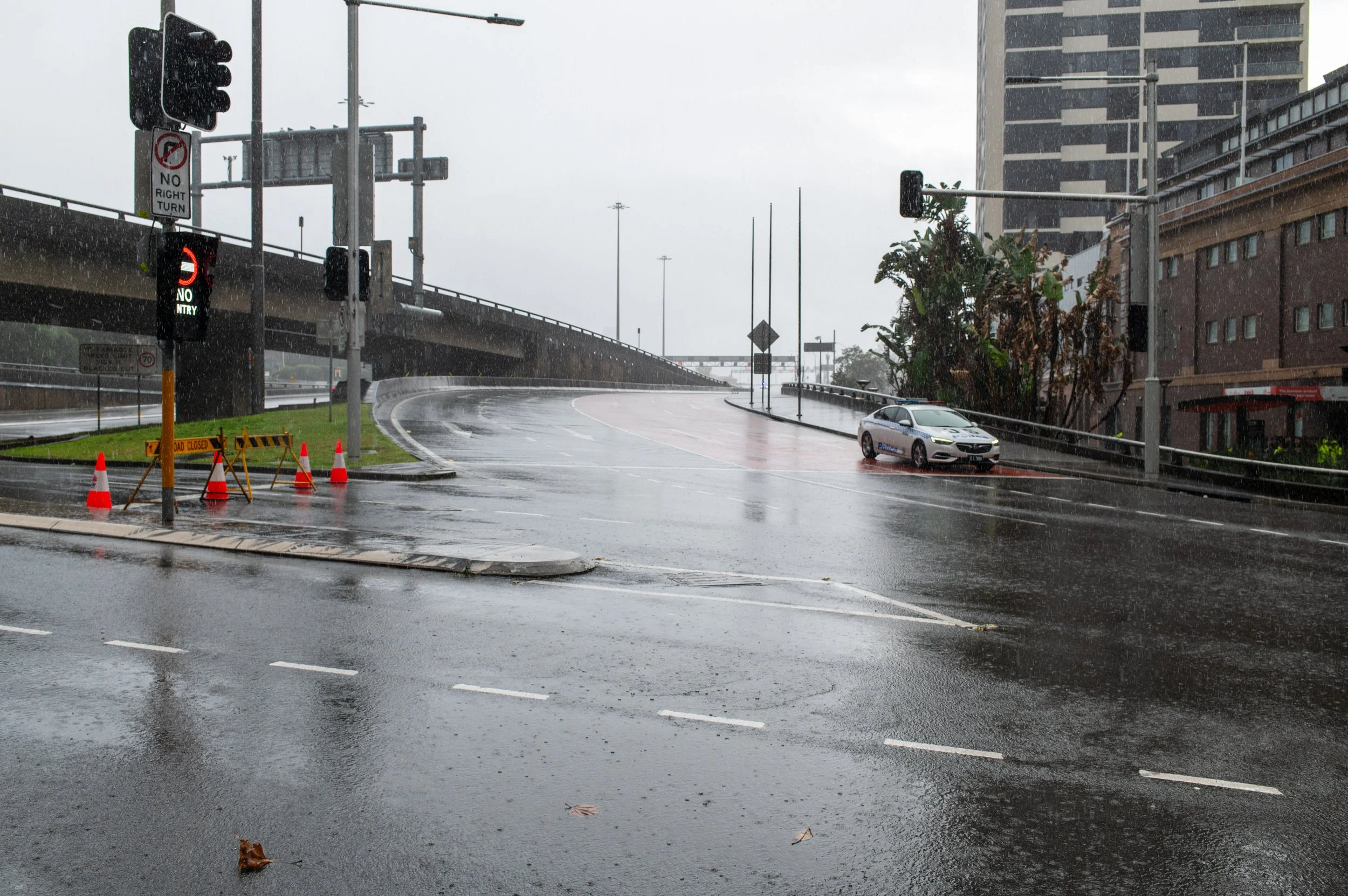 Rainy city street intersection with police car and street closure signs on wet pavement, traffic lights, and surrounding buildings.
