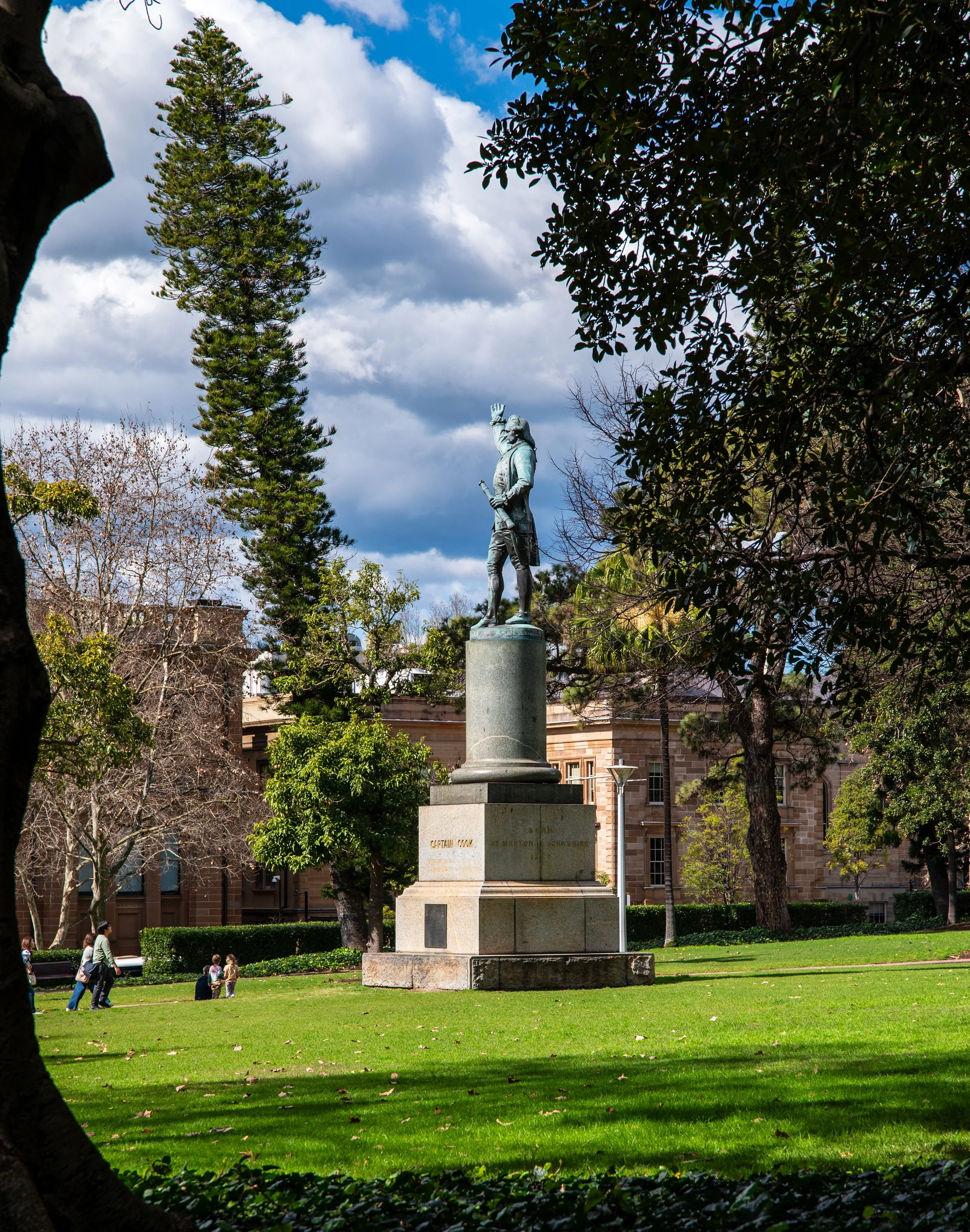 Statue of a man in a park, surrounded by trees and grass, with cloudy sky in the background.