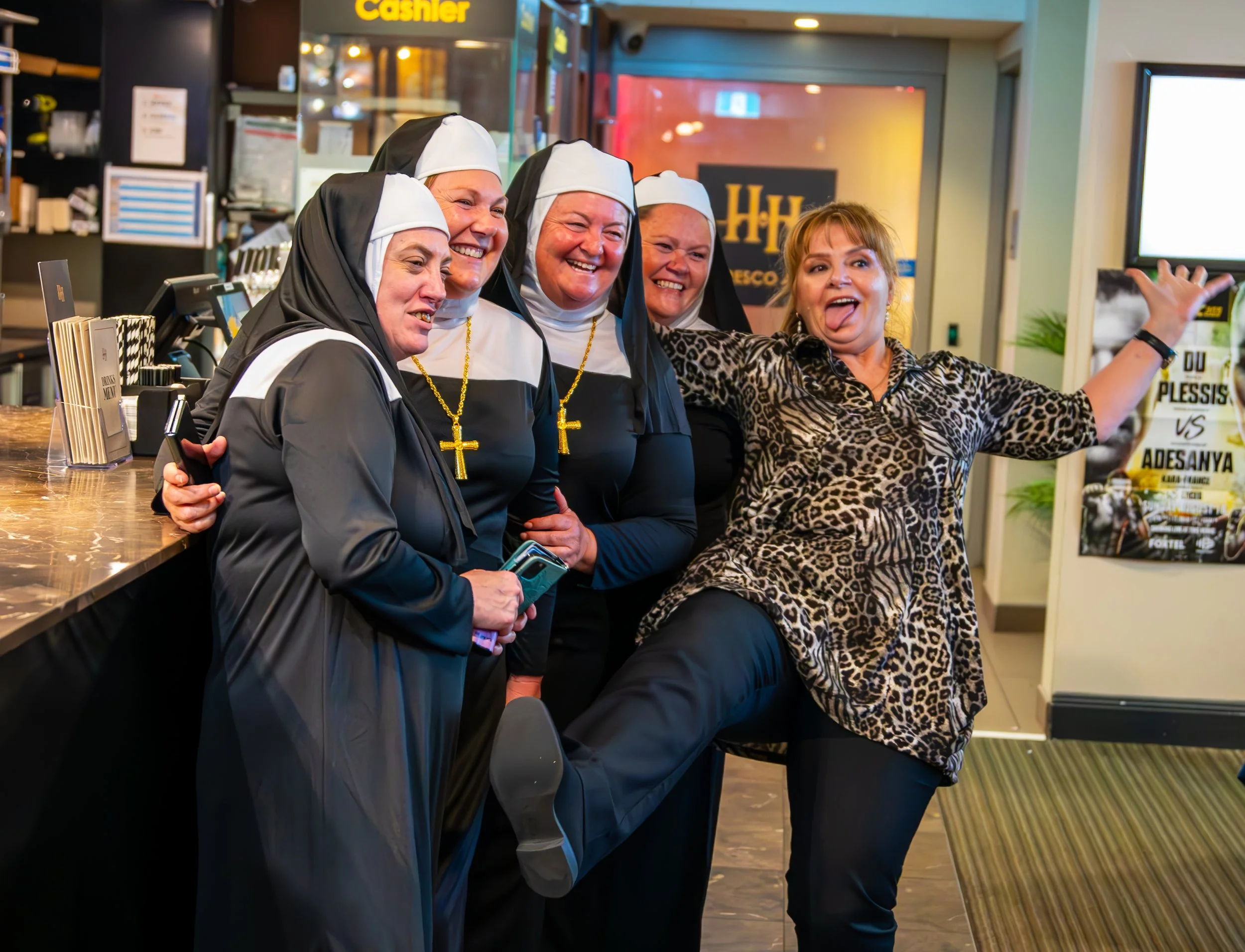 Four women dressed as nuns and a woman in a leopard print blouse posing happily and enthusiastically inside a restaurant or cafe, with one woman extending her arms in a lively gesture.