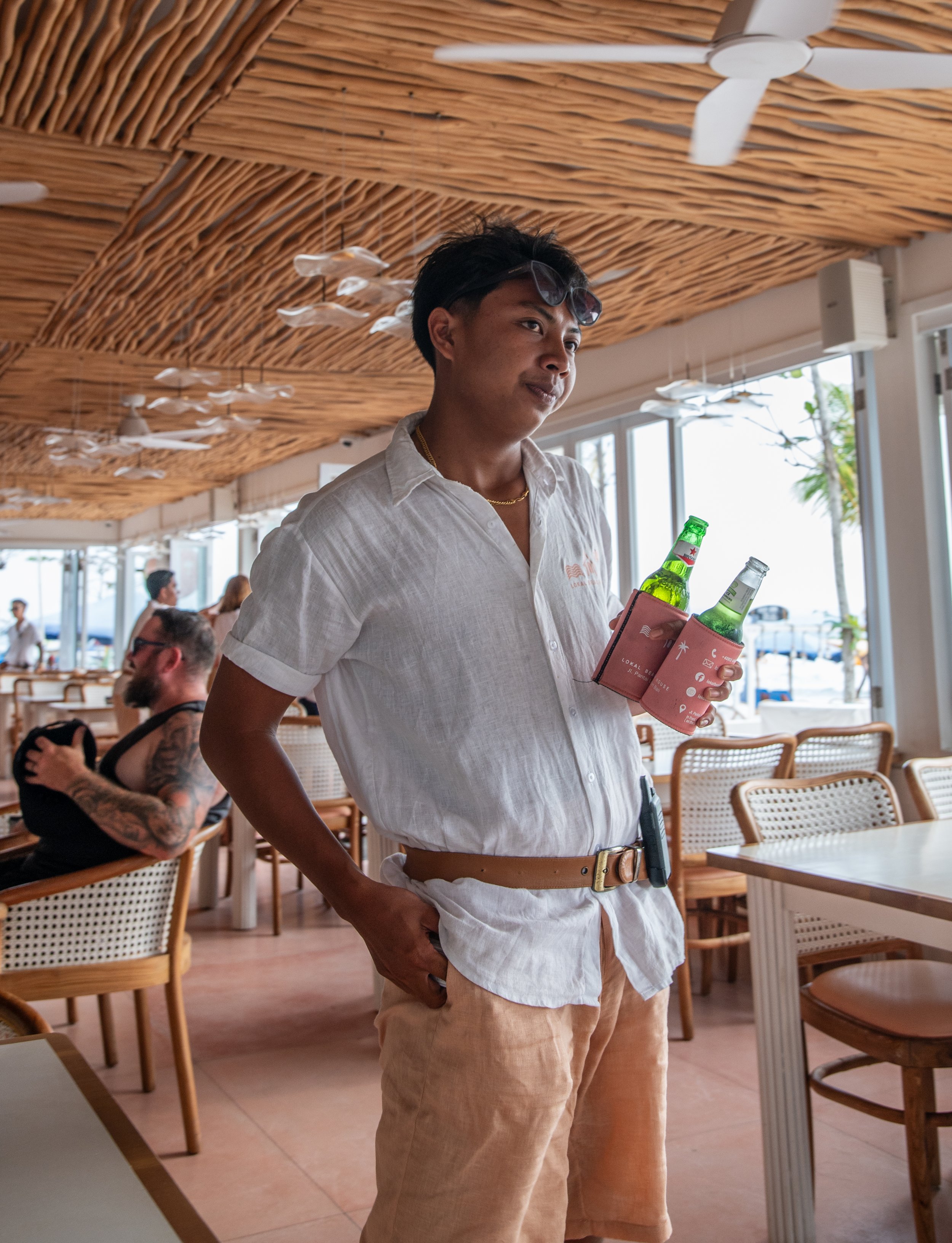 Young man holding several beer bottles and a pink wallet inside a restaurant or cafe.