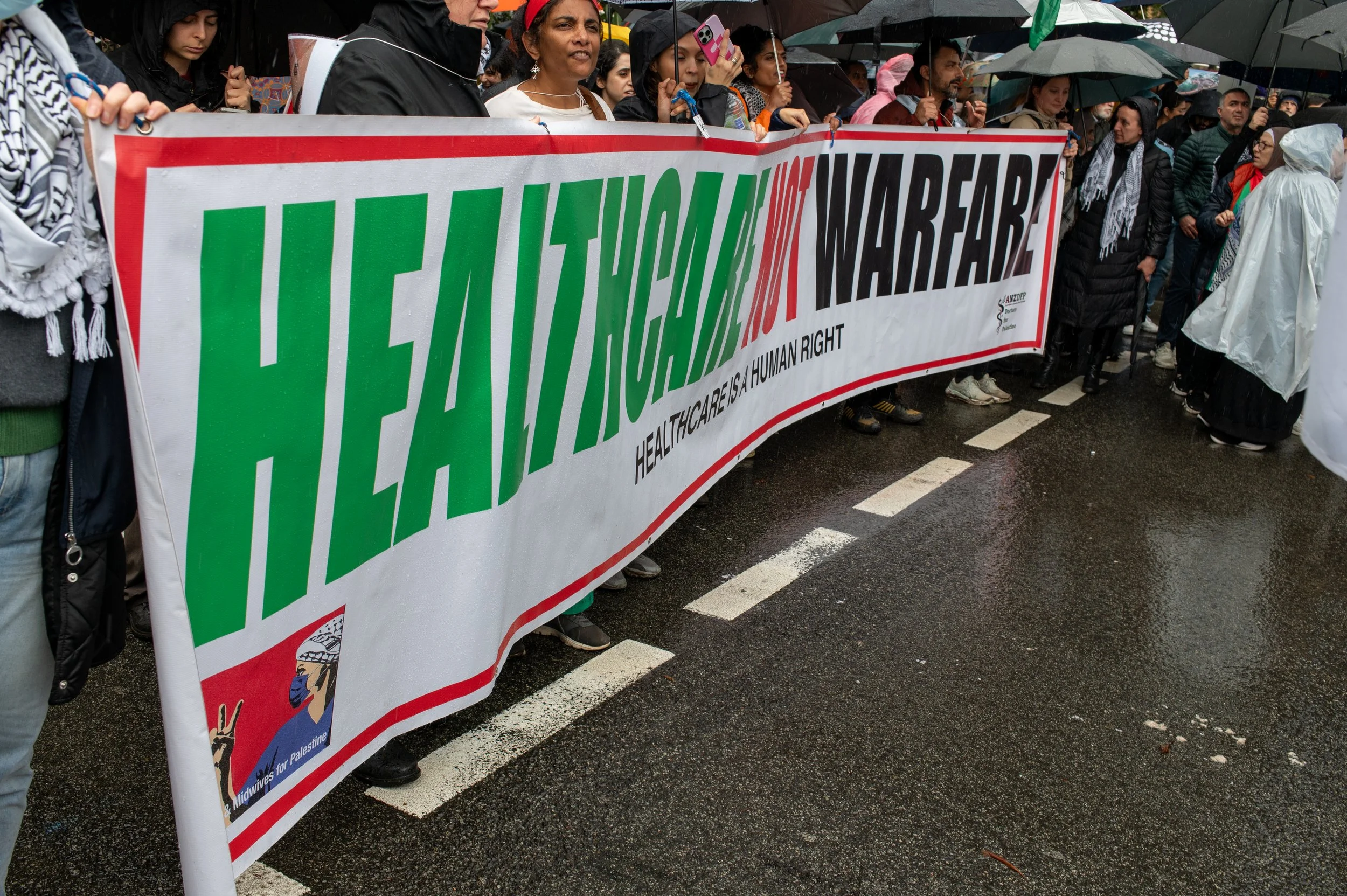 People holding a large protest banner that reads 'Healthcare is a Human Right' at an outdoor rally on a rainy day, with many attendees holding umbrellas.