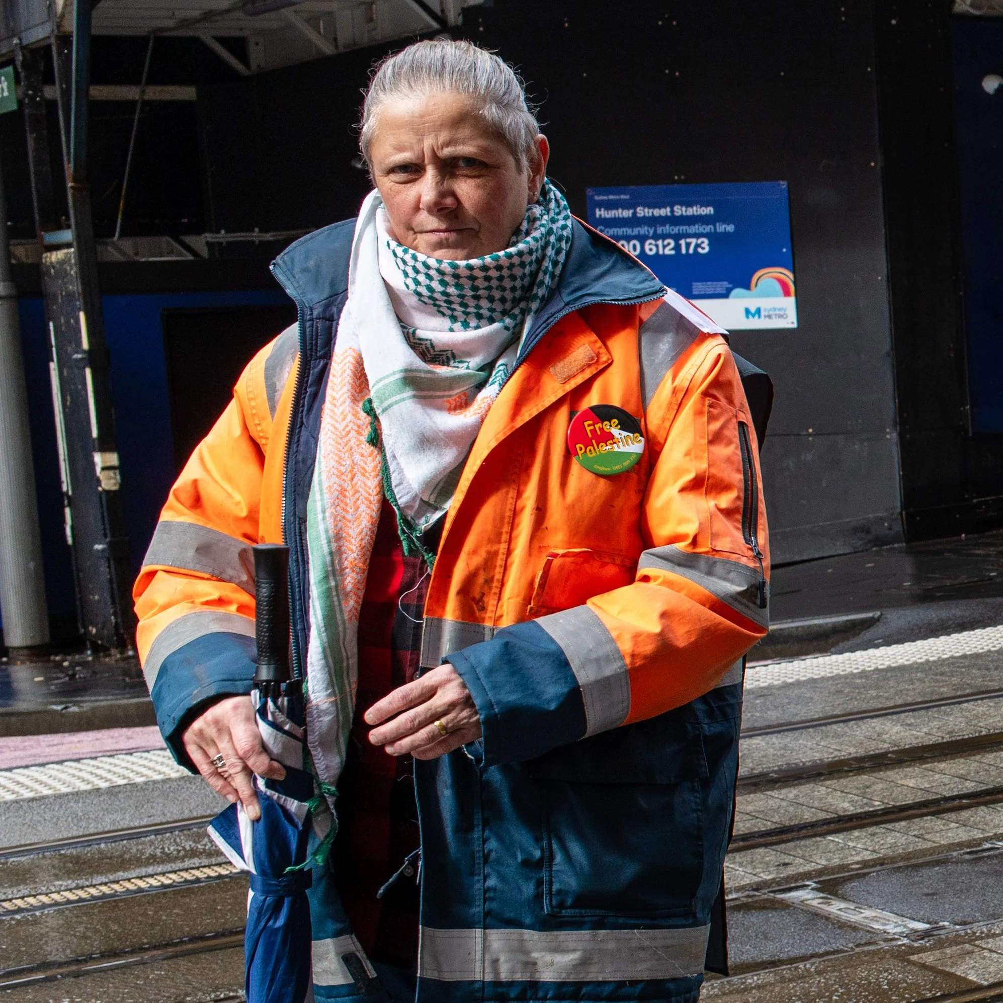Woman in orange and navy safety jacket holding an umbrella at a tram station with a sign that reads "Hunter Street Station" in the background.