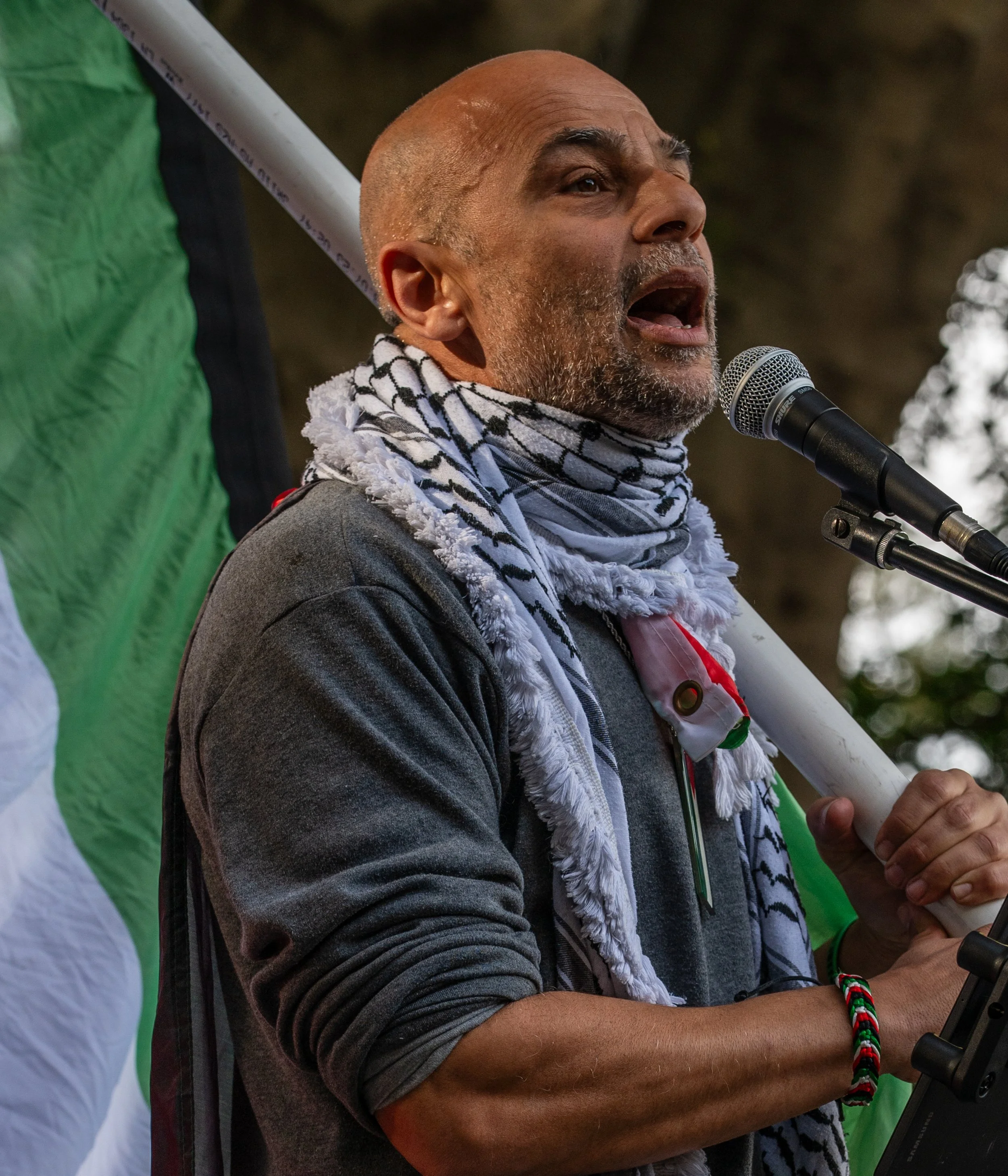 A man speaking into a microphone during a rally, with a black and white keffiyeh scarf around his neck and holding a Palestinian flag.