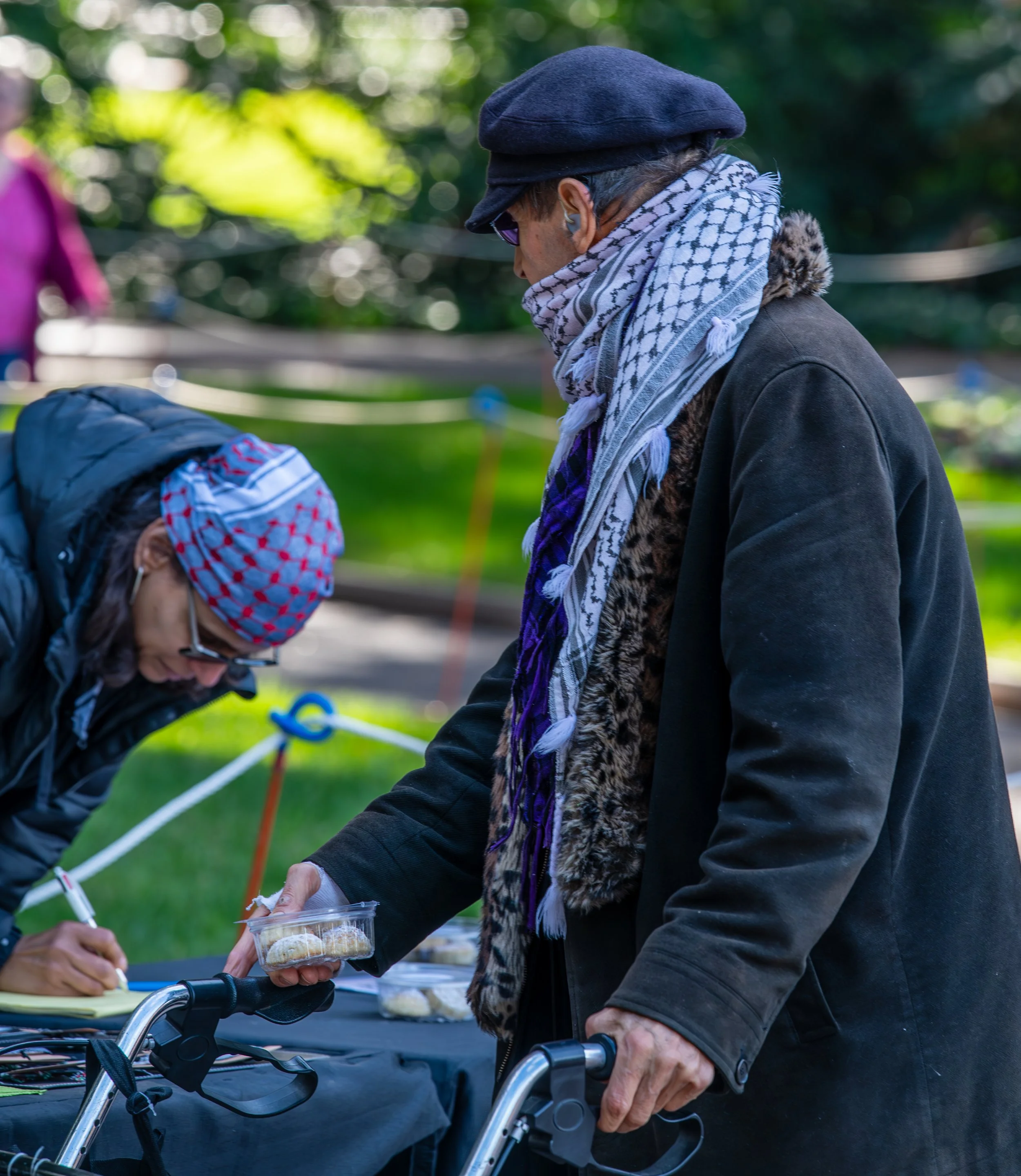 Two elderly women outdoors, one with a walker, exchanging a small food container, with a table and blurred greenery in the background.